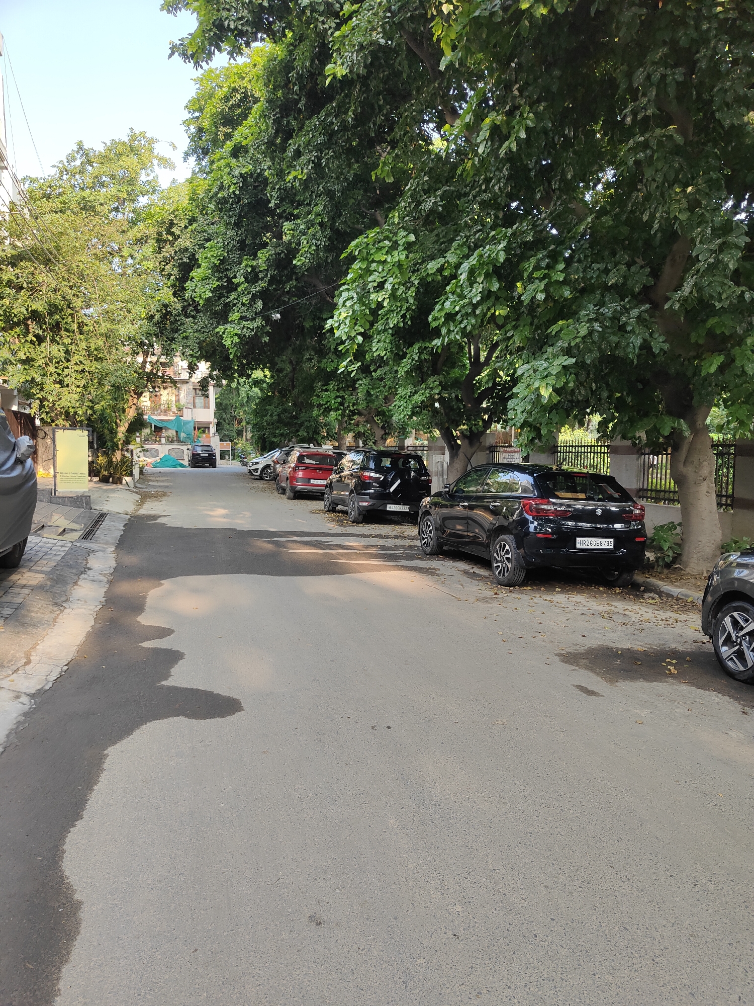 The image depicts a residential street in Gurgaon, India, lined with mature trees providing shade over parked cars. Several vehicles, including a black Maruti Suzuki Baleno with license plate HR26GE8735, a red SUV, and other black cars, are parked along the side of the asphalt road. A dark wet patch is visible on the road surface, indicating recent moisture. In the background, apartment buildings are visible, interspersed with greenery. The sky is clear and bright.