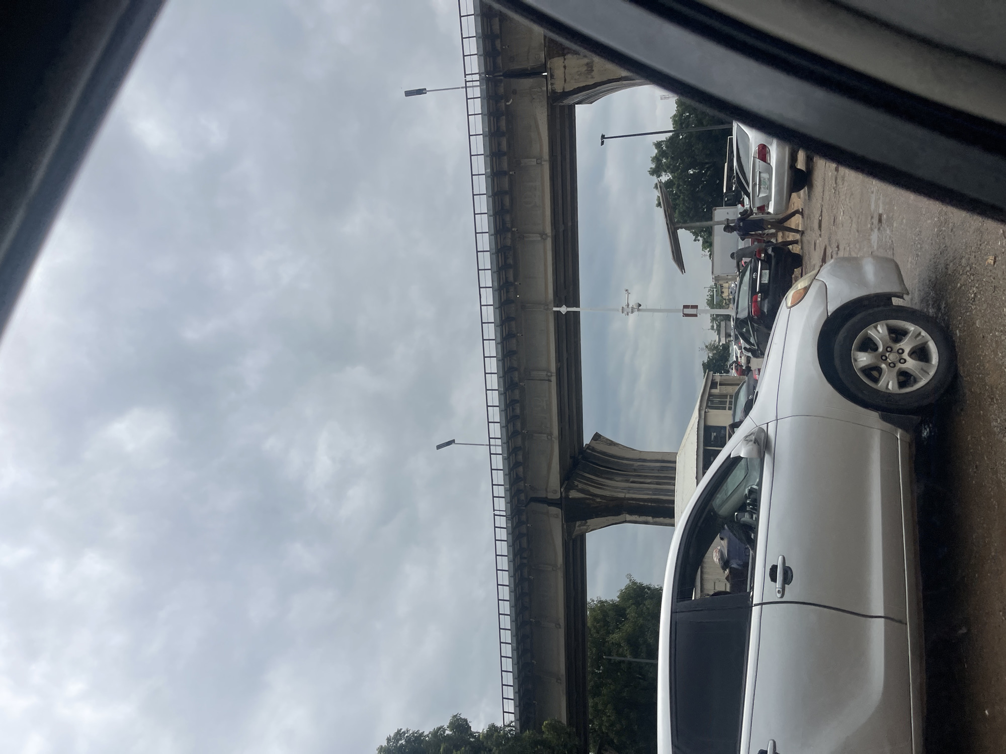 The image depicts a street scene in Gwaska, Nigeria, characterized by overcast skies and a concrete overpass spanning the road. In the foreground, the side of a silver car is visible, with its driver's side door open. Behind it, several other vehicles are present, including a black car and a silver sedan. Two individuals are standing near the black car, seemingly interacting. The ground appears to be unpaved and wet, suggesting recent rainfall or a drainage issue. Trees are visible in the background, and a building with a roof structure is situated behind the cars. The overall atmosphere suggests a busy urban environment possibly experiencing some traffic congestion.