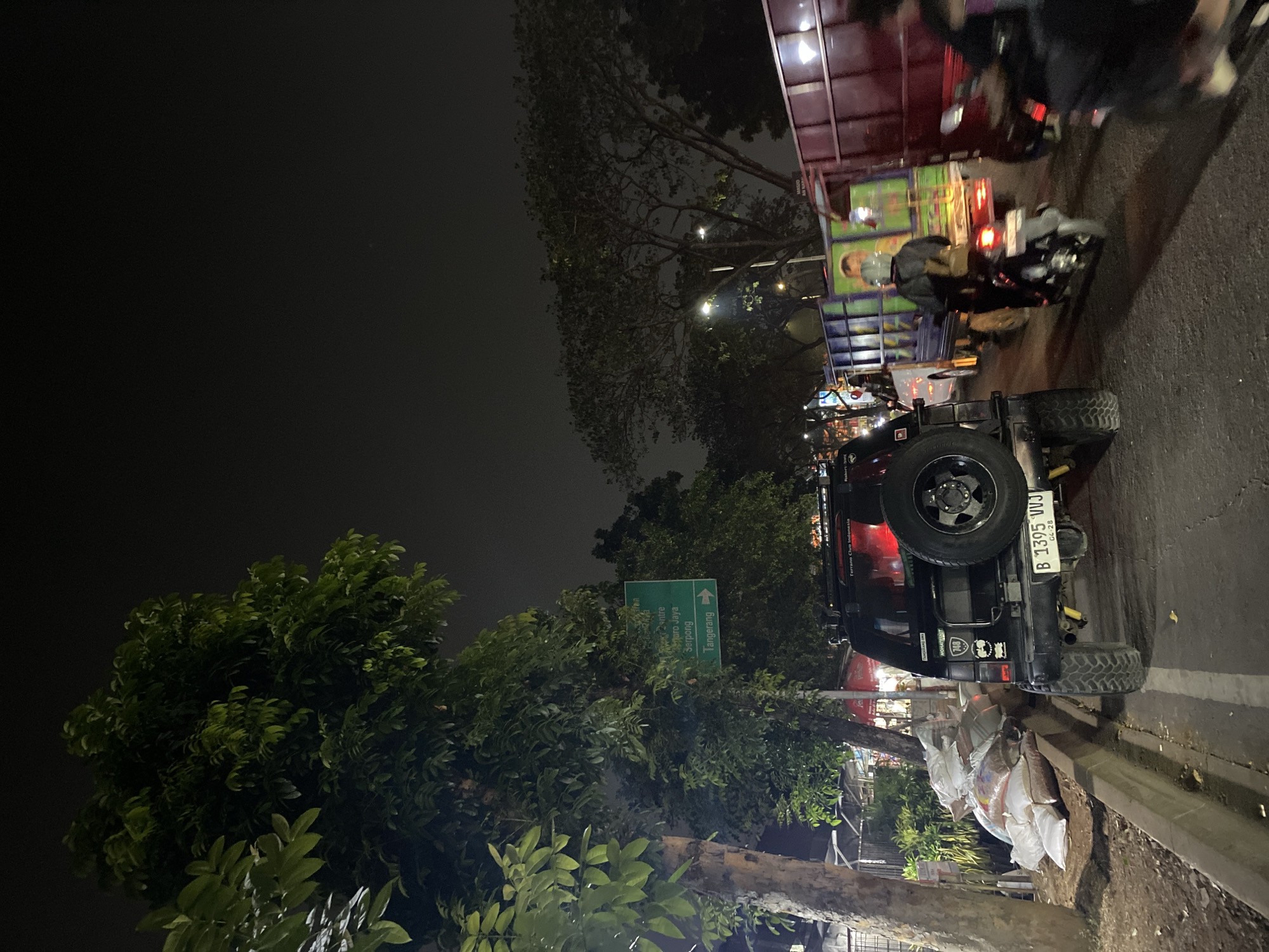In South Tangerang, Indonesia, a nighttime traffic scene unfolds on a city street. The foreground features the rear of a black SUV, its spare tire mounted externally. Adjacent to the SUV, a pile of large sacks, likely containing sand or grain, rests by the roadside. A green directional signpost is visible amidst lush foliage, indicating routes to "Centro Jaya Serpong" and "Tangerang" with an arrow pointing right. The mid-ground is characterized by heavy traffic congestion. A motorcycle with a rider wearing a helmet and a backpack navigates the gridlock. Behind it, a green and blue cargo truck is followed by a large red truck. The street is illuminated by vehicle headlights and streetlights, casting a dim glow on the surrounding trees and dark sky. The overall environment suggests a bustling urban area experiencing a traffic jam at night.