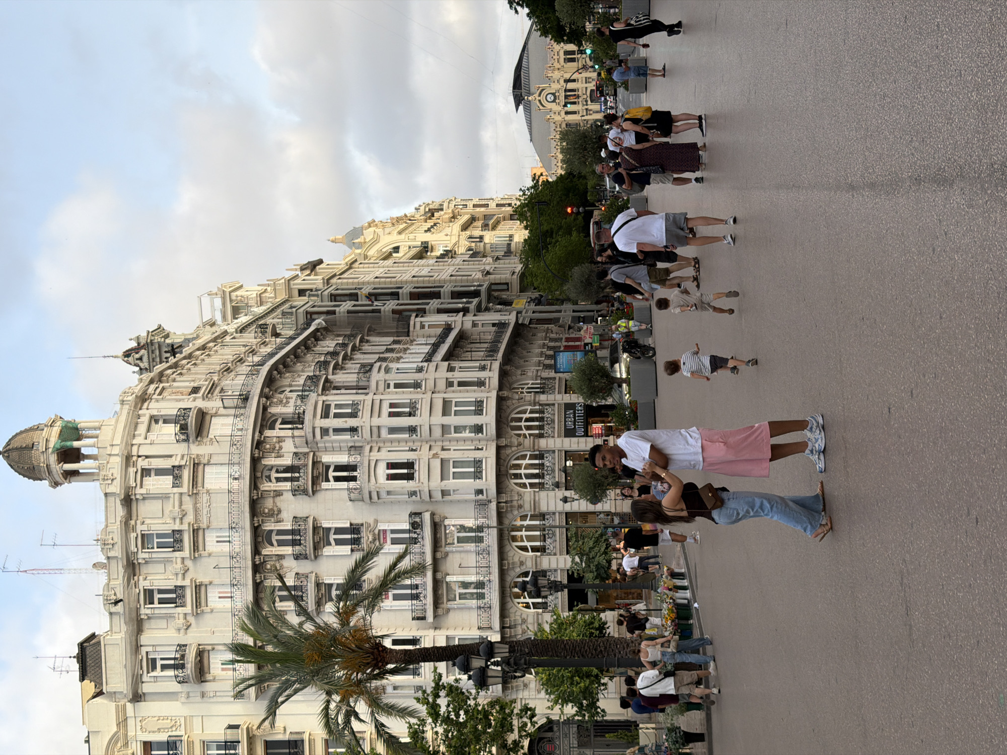 The image depicts a bustling city street scene with people walking and interacting. Dominating the background are ornate, multi-story buildings with detailed facades, balconies, and numerous windows, suggesting an older, European architectural style. A tall palm tree stands prominently in the left foreground. In the mid-ground, a diverse group of people are visible, including couples, families with children, and individuals. Some are walking, others are conversing, and a couple in the foreground is engaged in a close interaction. Various objects are present, such as street lamps, planters with flowers, and signs for businesses like "URBAN OUTFITTERS". The ground is paved, and the sky is partly cloudy. The overall atmosphere suggests a public gathering space within a city.