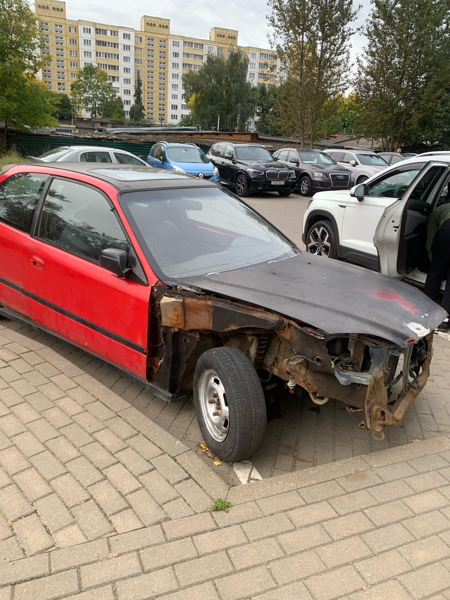 A red, two-door coupe, with its hood and front bumper removed, revealing significant rust and damage, is parked on a brick-paved lot. The car's interior is visible, including the dashboard and a portion of the passenger seat. Several other vehicles, including a white SUV with its driver's door open, a dark blue hatchback, a black SUV, and a gray SUV, are parked in the background. A tall, yellow apartment building with multiple windows and a gray, overcast sky are visible behind the cars. The scene appears to be a parking lot in an urban environment, possibly a residential area in Minsk, Belarus, given the architecture. There is no visible interaction between people and the red coupe.