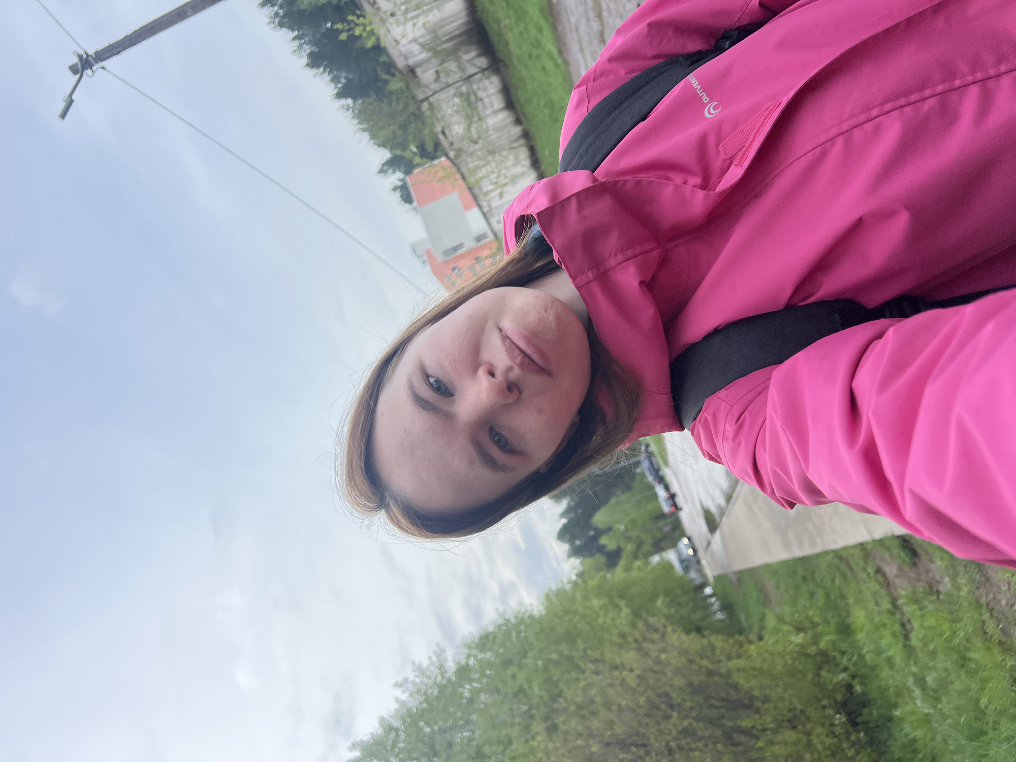 A young woman wearing a pink jacket and black backpack is taking a selfie outdoors on what appears to be an overcast day. In the background are trees and foliage, a paved pathway or road, some parked cars, and a building with a distinct red roof. A utility pole and wires are visible in the top left corner of the frame. Based on the available visual information, the woman appears to be in a park or green space in Chaikovsky, Russia, due to the presence of the red-roofed building visible in the background.