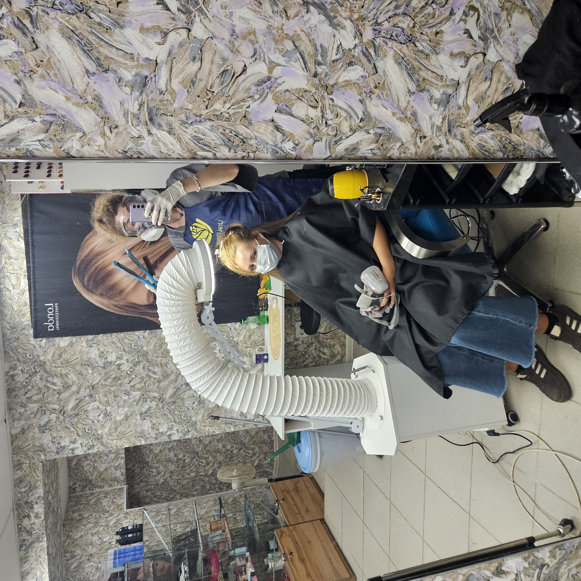 A young woman with blonde hair, wearing a black cape and blue jeans, sits in a salon chair with a mask on. She holds a respirator mask in her lap. A large, white, flexible hose with a light is positioned above her head. A masked and gloved stylist stands behind her, holding a phone and a hair straightener. The salon features patterned wallpaper and shelves stocked with hair products.