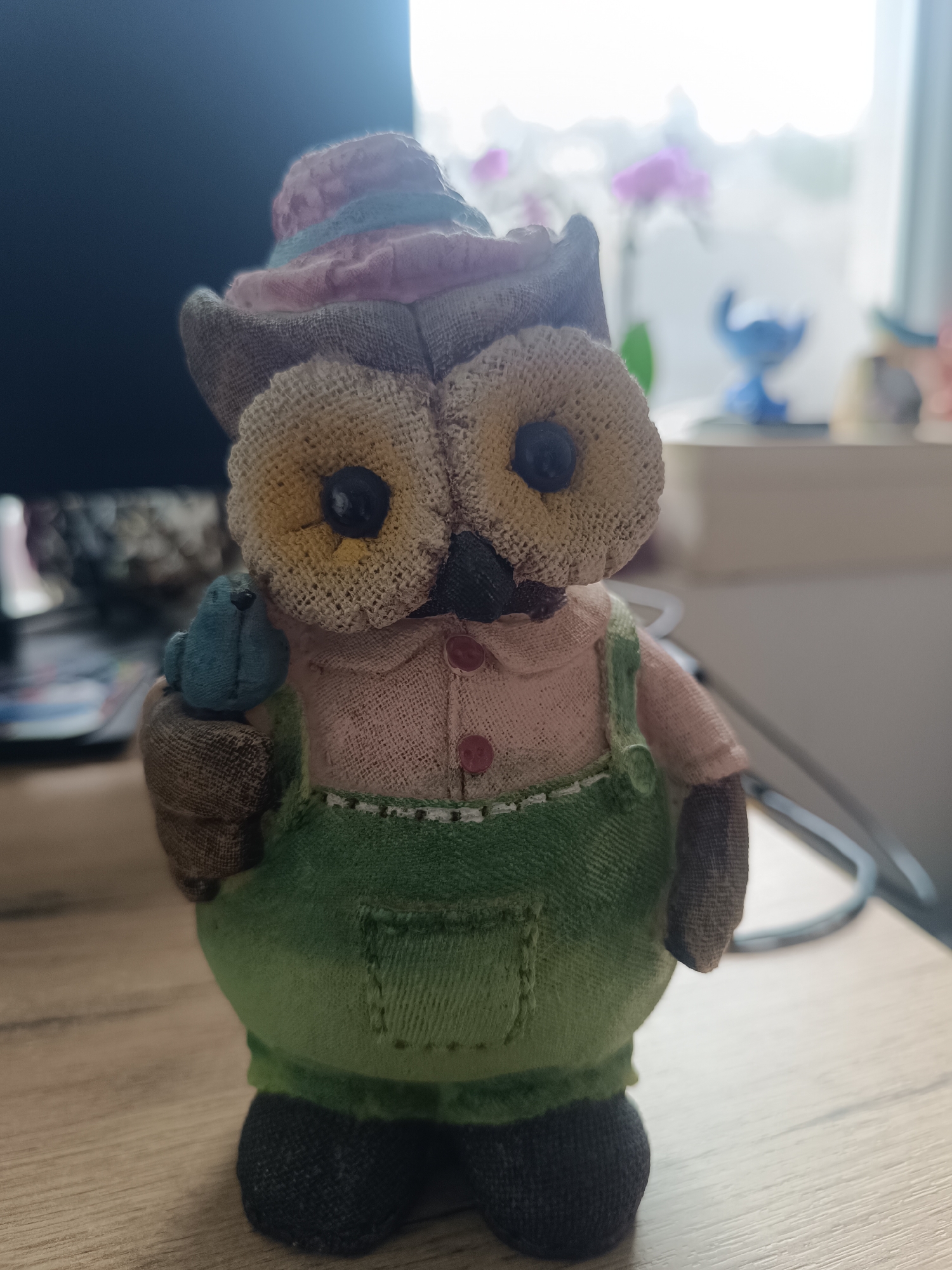A fabric owl figurine stands centered on a light wooden surface in an indoor setting. The figurine features a rounded body, wearing a pink and purple striped hat with a light blue band, a light pink shirt with two visible reddish-brown buttons, and green overalls with a front pocket. Its face is made of a coarse, light-brown fabric, with two black button eyes and a black pointed beak. A small, light blue fabric bird is held in its left (viewer's right) brown fabric arm. The background to the left is dark and blurred, suggesting an electronic screen or monitor. To the right, a brightly lit and heavily blurred window is visible, with indistinct shapes including a purple orchid flower and a small blue figurine resembling 'Stitch' on a white shelf or sill. This scene is located within the city of Varna, Bulgaria.