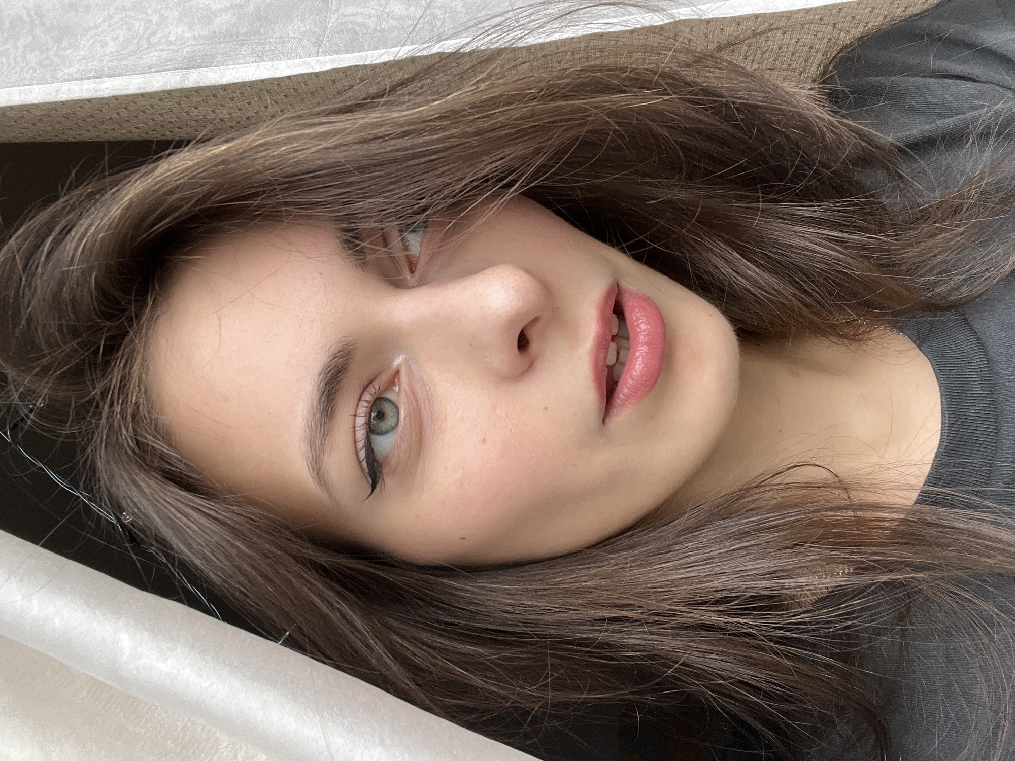 A young woman with brown hair and green eyes is depicted in a close-up, slightly tilted selfie. She has fair skin and is wearing subtle makeup, including eyeliner and pink lipstick. Her hair is styled loosely around her face. A small portion of what appears to be a grey or black garment is visible in the upper right corner. The background consists of a white textured surface and a dark, undefined area. The location in the city of Krasnoyarsk, Russia is not determinable from the visual information.