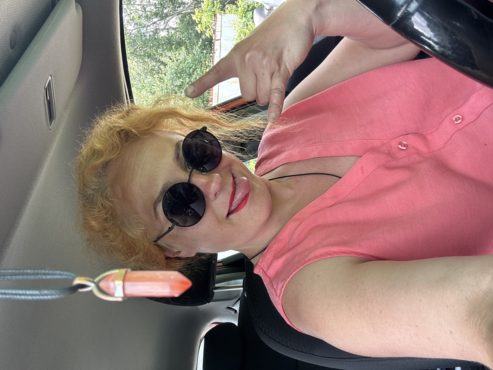 A fair-skinned woman with short, curly blonde hair is in the driver's seat of a car, wearing large, round, black sunglasses and a bright pink sleeveless V-neck shirt. She is sticking her tongue out playfully and making the "shaka" hand gesture with her right hand, while a pink crystal pendant hangs from the rearview mirror. The car's interior is visible, with a black headrest and seat. Outside the car, greenery and a white structure are visible through the windows, suggesting a somewhat rural or suburban setting, possibly on the outskirts of Chaikovsky, Russia. The lighting indicates it is daytime.