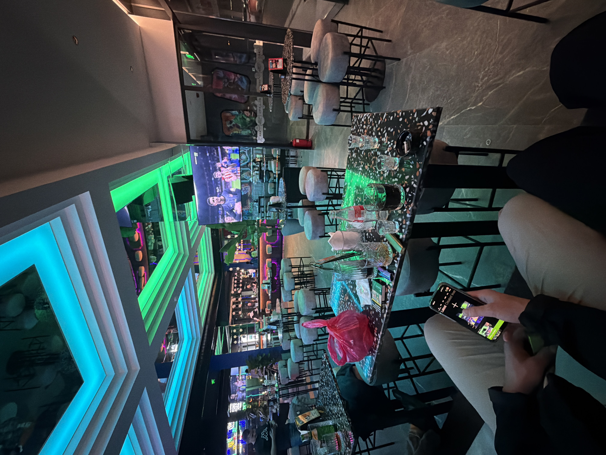 A tilted, long-exposure photograph shows the interior of a bar or restaurant, likely in Varna, Bulgaria given the information provided. A person sits at a table with a distinctive terrazzo-like top, holding a smartphone with a bright screen. The phone displays what appears to be a game or app with colorful, abstract graphics. There are several glasses on the table and what looks like a red plastic bag. In the background, there are numerous tables and bar stools, along with shelves displaying bottles. The bar itself is lit with green and blue neon lighting, creating a modern and lively atmosphere. A large screen is visible in the background, displaying a soccer or sports match. The overall ambiance suggests a nightlife setting.