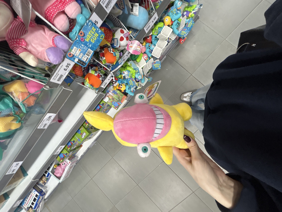 The image captures a first-person perspective of a person holding a distinct yellow and pink plush toy in what appears to be the toy aisle of a retail store. The plush toy, which is the central focus, is primarily yellow with a large pink section covering its face, featuring a wide, stitched smile with visible white teeth. It has two prominent, light blue googly eyes, one positioned above the pink facial area and another below. The person's right hand, with dark manicured nails, holds the toy.
The background reveals multiple metal-framed shelves stocked with various colorful toys. On the upper shelves to the left, pink plush toys are visible, along with multiple blue plastic "WATER GUN" packages. Further along the shelves, a variety of other toys are displayed, including colorful plastic items, some with eyes, and mesh bags containing smaller, brightly colored objects, possibly balls or toys. White pricing labels are affixed to the shelves, displaying prices such as "6.95", "5", "3.99", and "2.99". One visible label specifically mentions "WATER GUN" and the "CE" mark. Another small, pink tag partially visible behind other toys reads "SQUEAKY TOYS".
Below the shelves, the person's lower body is visible, dressed in dark long-sleeved clothing (possibly a hoodie or jacket) and light blue jeans. Their feet, clad in white sneakers, are also seen on the light grey tiled floor of the store. The lighting suggests an indoor retail environment. The image is taken from a slightly overhead angle and is rotated, giving a dynamic, casual snapshot feel.