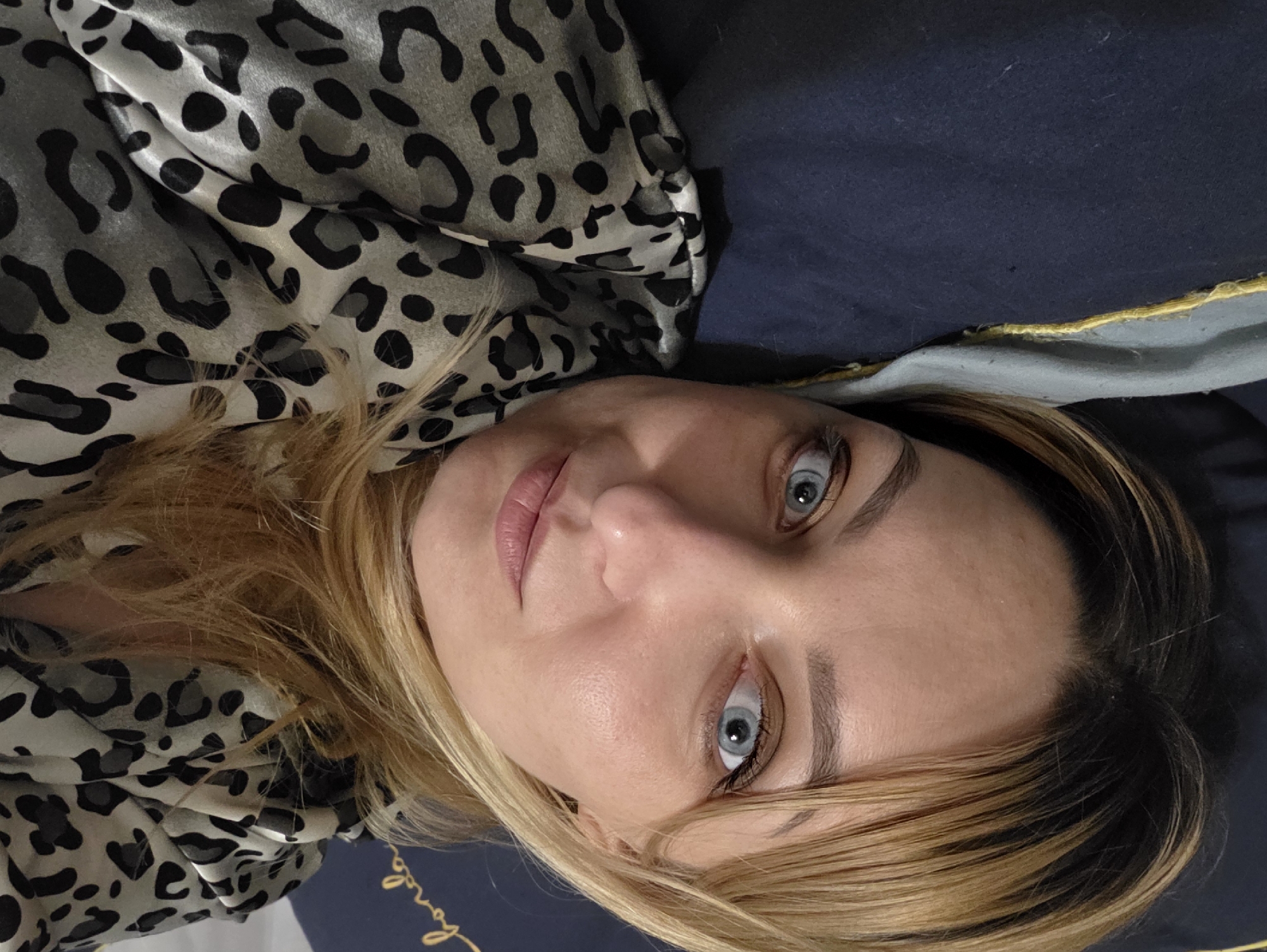 A female subject with light skin, blonde to light brown hair with darker roots, and blue-grey eyes is depicted in a close-up, horizontal orientation, looking directly at the viewer. She is situated on what appears to be a bed in a domestic indoor setting, specifically in Minsk, Belarus. Her head rests on a dark blue pillow or duvet, which features a distinct light grey-blue fabric border with a bright yellow stitched piping. She is wearing a garment with a grey and black leopard-print pattern, which appears to be made of a silky or satin-like material. Her facial features include groomed eyebrows, visible mascara on her eyelashes, and a light pink lip color. The overall impression is one of relaxed repose.