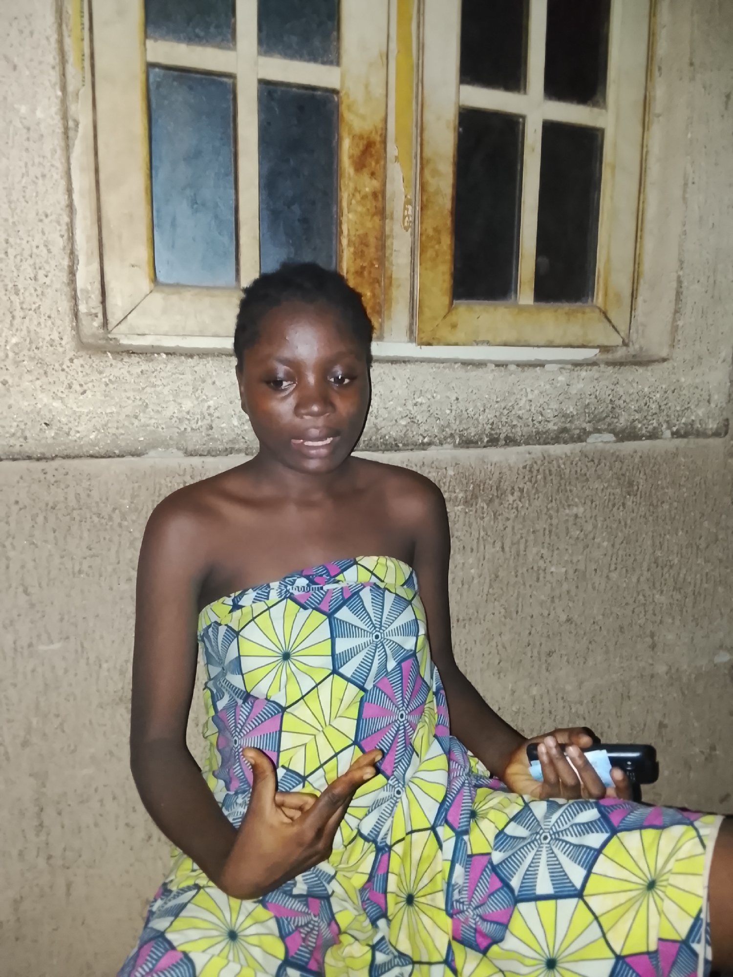 A young woman is depicted sitting or leaning against a textured, light-colored wall, possibly made of concrete or stucco, in what appears to be a domestic setting in Akwanga, Nigeria. Behind her, an old-fashioned window with multiple dark panes is visible, suggesting either nighttime or low light conditions. The window frame is aged and discolored.
The woman has dark skin and short, dark hair. She is wearing a strapless garment or wrap featuring a vibrant, geometric pattern with yellow, pink, blue, and white colors. Her expression is neutral, and she is looking slightly to her right, towards the camera.
Her left hand is positioned in a distinct gesture, with the thumb and pinky fingers extended and the middle two fingers curled. In her right hand, she holds a dark-colored smartphone, around which a light-colored material, possibly paper or tissue, is wrapped. The lighting in the scene appears to be artificial and somewhat uneven, creating shadows and highlights on the subject and background. There is no visible text in the image.