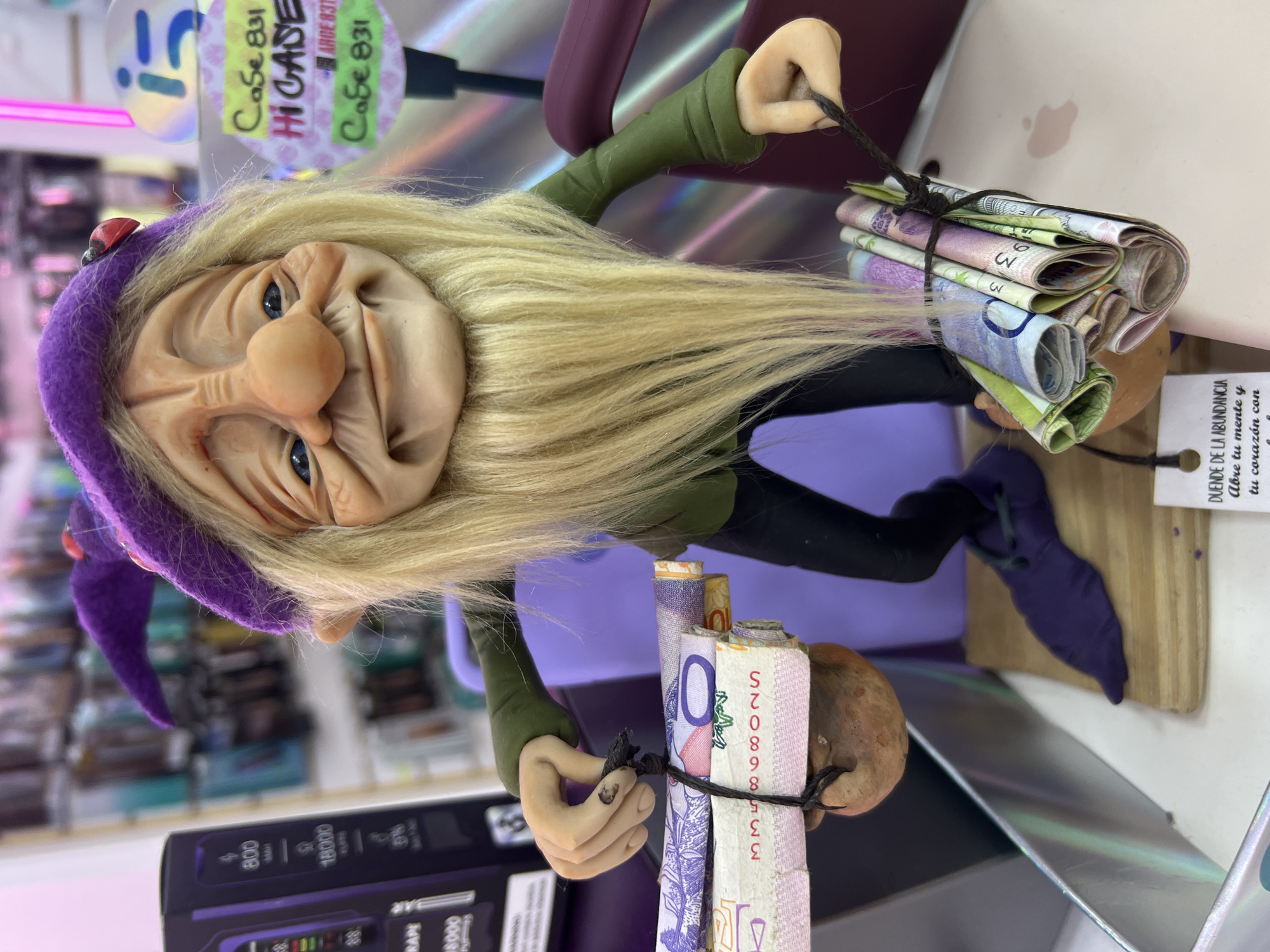The image shows a doll-like figure of an elf or gnome, dressed in green and black with a purple hat and holding a bundle of Argentinian pesos tied together with black string. The figure has light skin, blue eyes, and a long white beard. The figure is positioned in front of a purple surface, with parts of various products visible in the background, including what appears to be a cell phone case. A small white tag with text in Spanish is attached to the doll. A sticker with the text "Case 831 HI CASE ARGE 83T Саб€ 831" is visible behind the doll. A product package with the text "800 MAH 18000" can also be seen. The currency held by the doll is Argentine pesos, and the Spanish text on the tag and the product package likely indicates that this is a product being sold in Buenos Aires, Argentina.