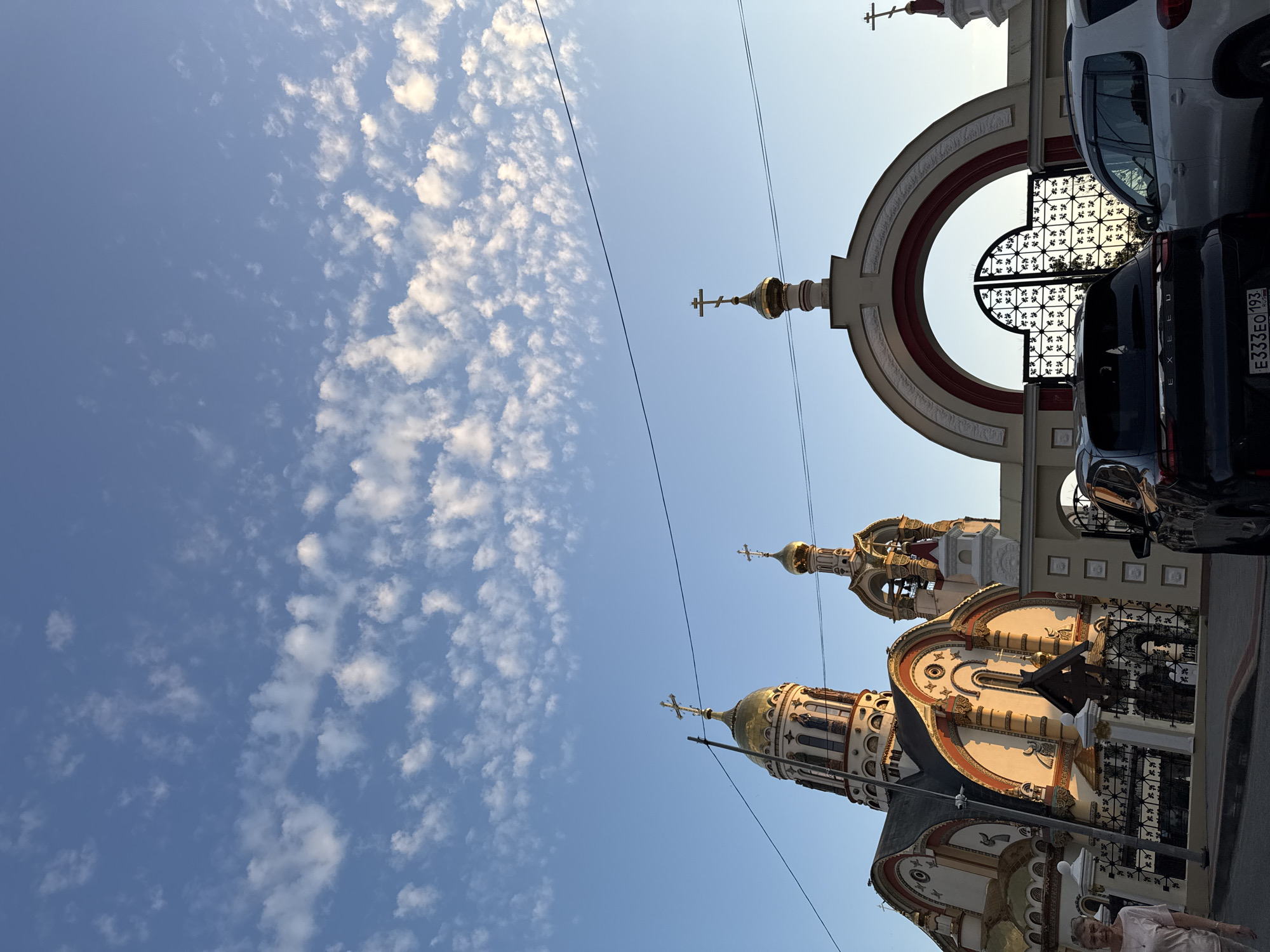 The image depicts the Church of the Icon of the Mother of God "The Joy of All Who Sorrow" in Sochi, Russia. The church is characterized by its golden domes and ornate architectural details, including arches and crosses. The sky above is a clear blue with scattered white clouds. In the foreground, two cars are parked in front of an arched gate with intricate metalwork. A woman is walking on the sidewalk to the left of the frame, and a black SUV with a license plate reading "E333EO 193 RUS" is partially visible. The scene appears to be captured during the golden hour, with soft sunlight illuminating the church's facade.