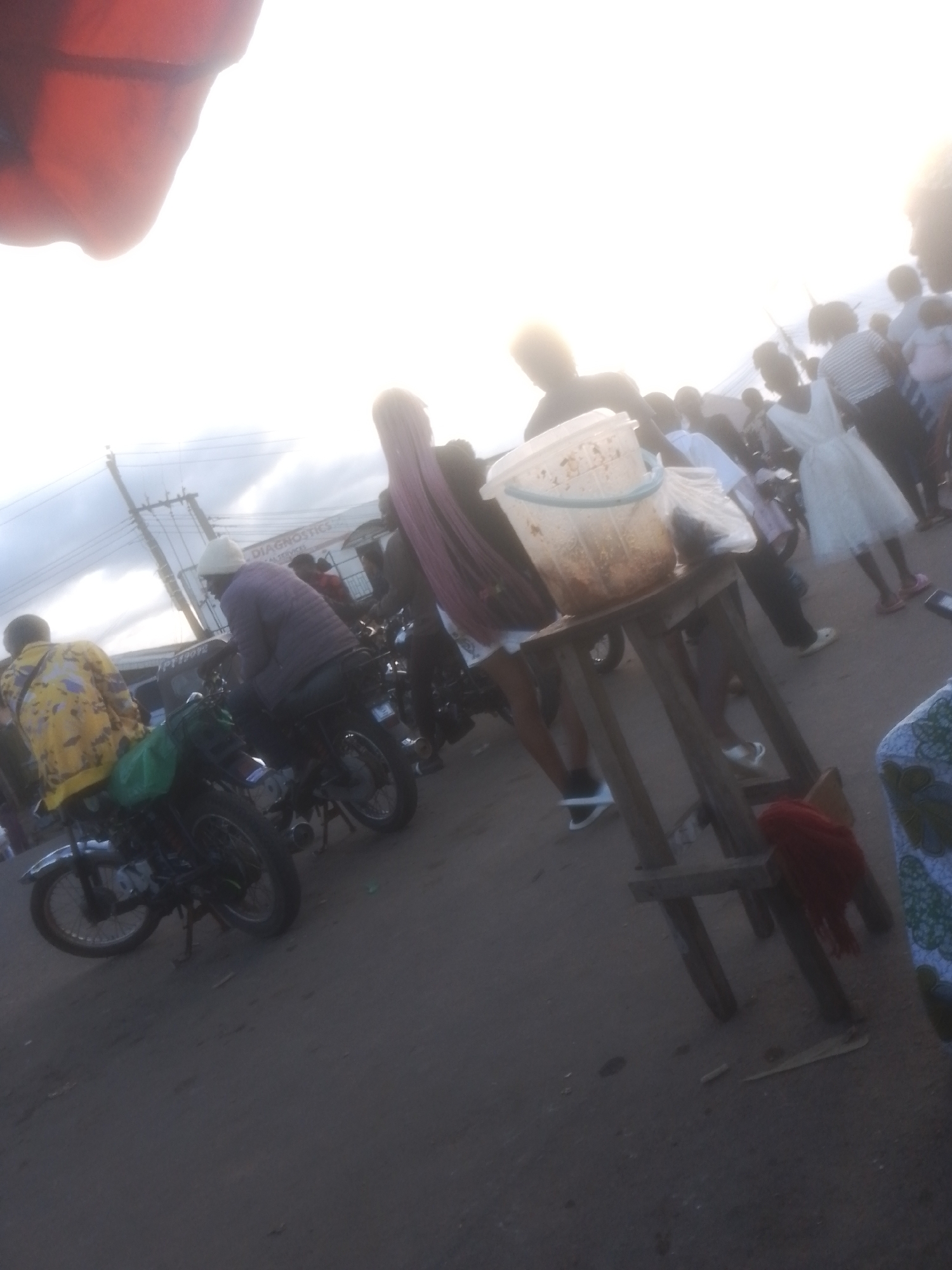In Gwong, Nigeria, a street scene unfolds with a bright, hazy sky. Several motorcycles are parked on a dusty, uneven surface. On the left, a person in a yellow jacket with a green bag on the back of a motorcycle is visible from behind. To their right, another individual on a motorcycle wears a purple jacket and a light-colored beanie. In the center, a wooden stand holds a large plastic bucket filled with what appears to be food, possibly snacks or ingredients. A person with long, dark hair is standing near the stand, wearing a white skirt. Further to the right, a woman in a white dress is visible, and other people are scattered throughout the background, some appearing to be waiting or observing. A building with the sign "DIAGNOSTICS" is present in the background, suggesting a commercial area. The overall impression is one of a busy, informal marketplace or gathering.