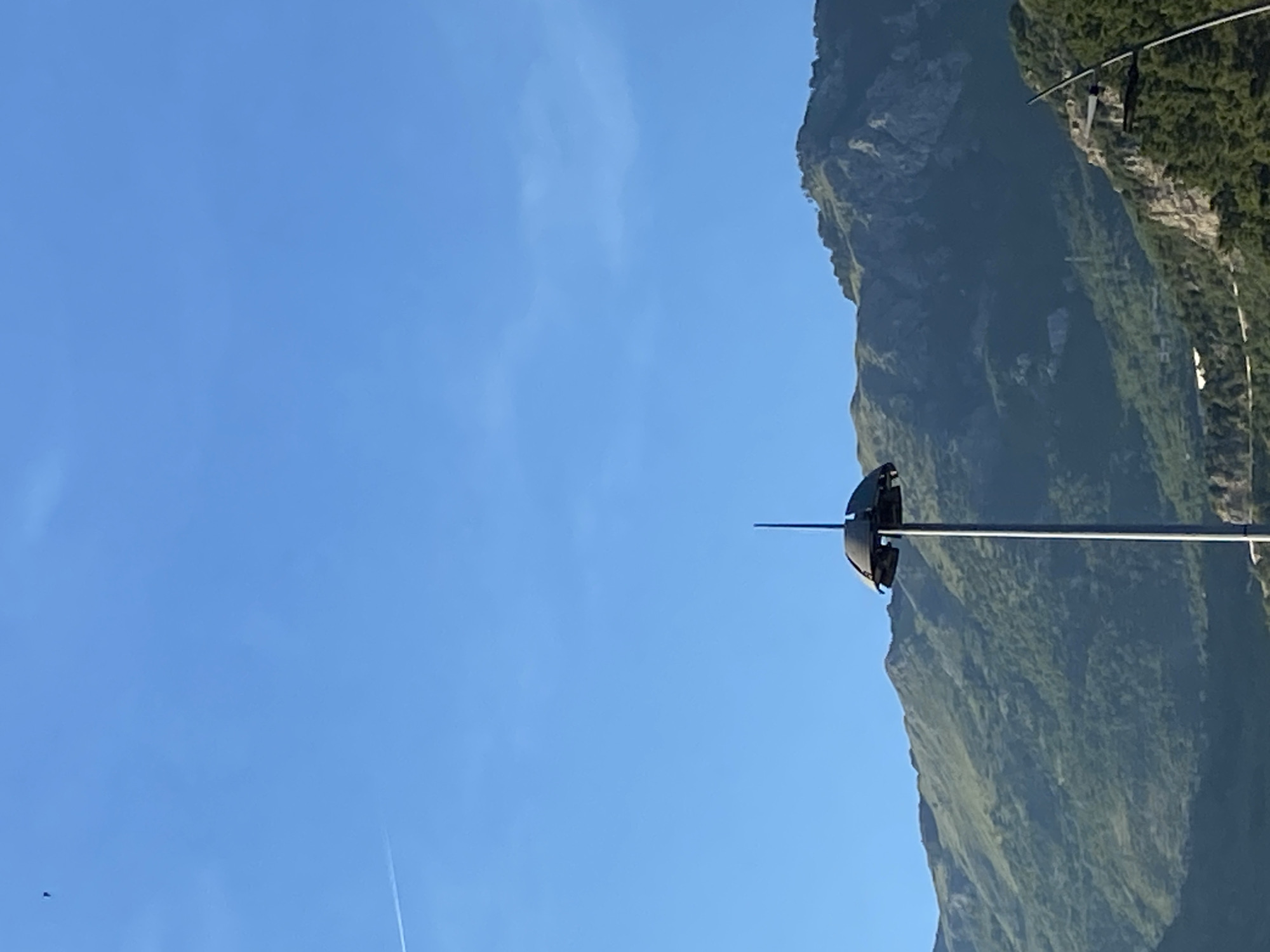 The image shows a tall, slender pole with a dome-shaped structure at the top, resembling an umbrella, against a clear blue sky. The pole is metallic and extends upwards, with a pointed finial at the very top of the dome. In the background, there are lush green mountains with rocky outcrops. A faint white contrail from an aircraft is visible in the upper left quadrant of the sky. The scene appears to be outdoors, possibly in a mountainous region. The specific location in Budva, Montenegro, is not identifiable from the visual information alone.
