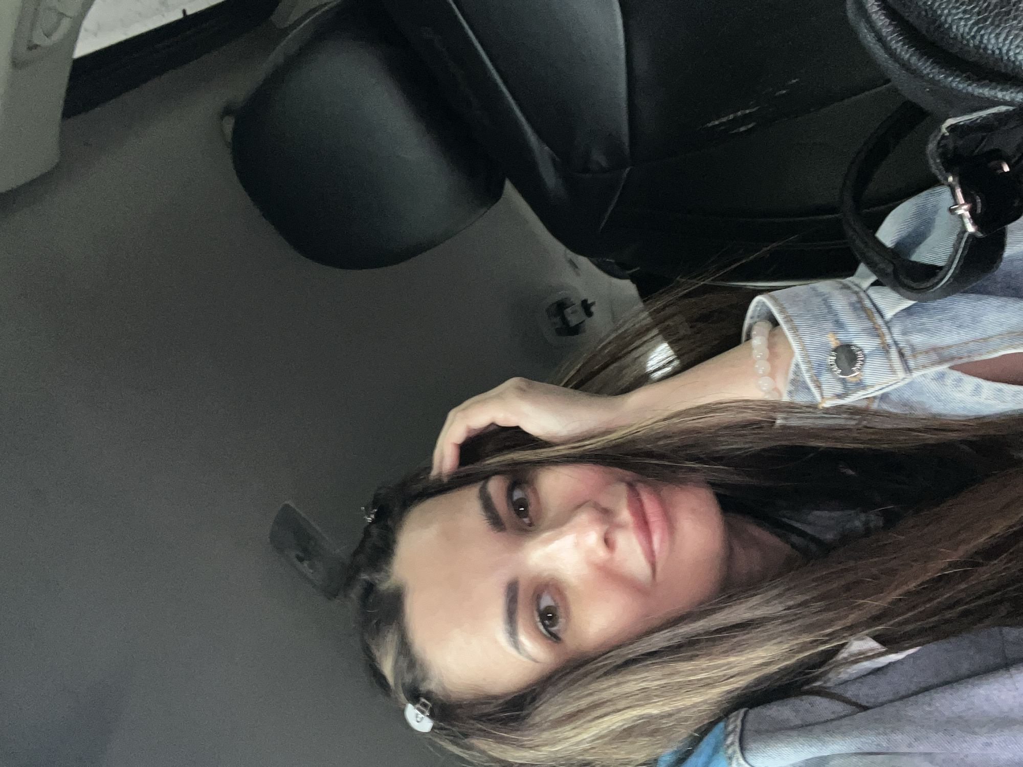 A woman with long brown hair, wearing a denim jacket and bracelet, is sitting in a car. The car seat and steering wheel are visible in the background. The location is Unknown, Unknown.