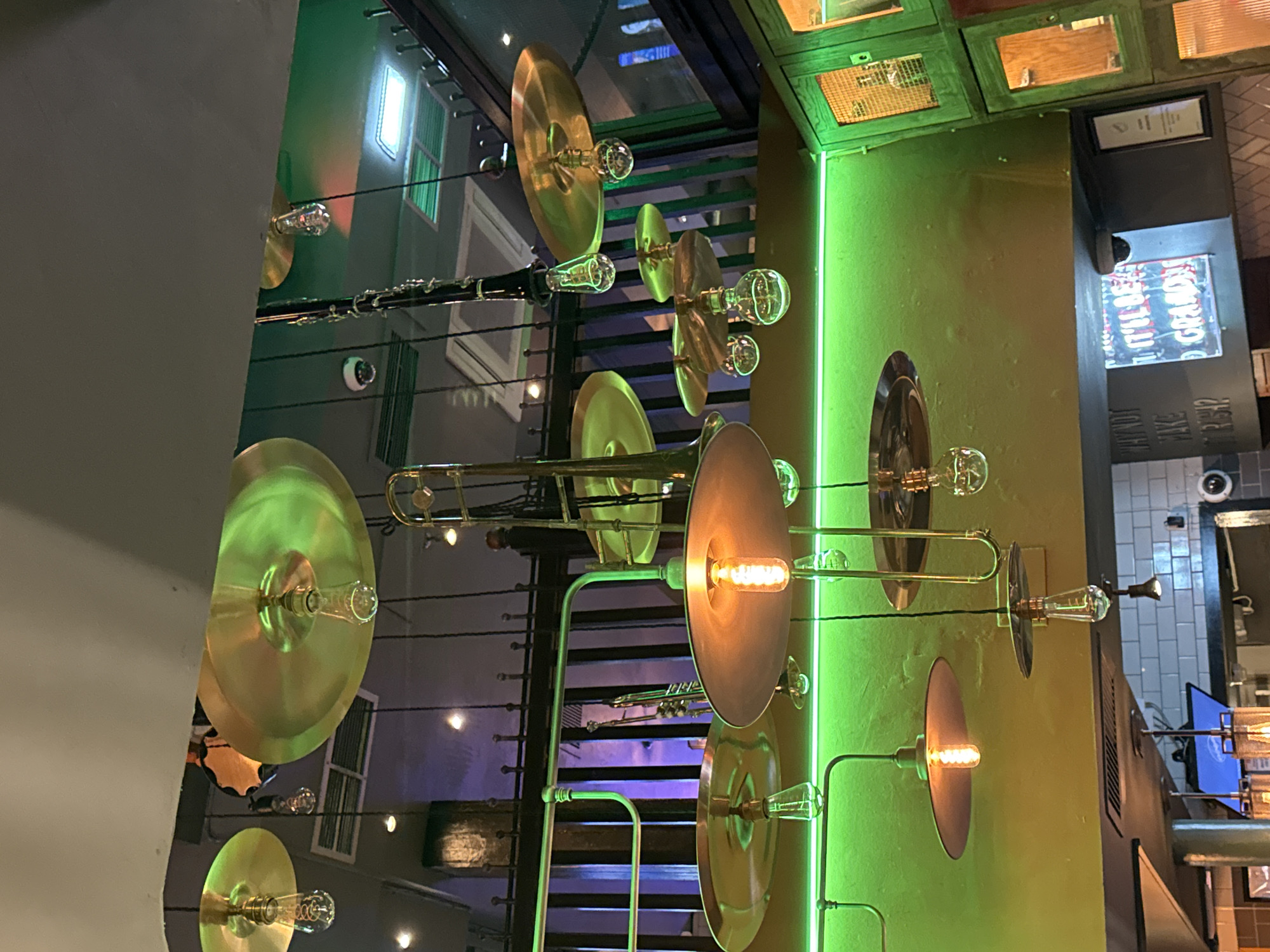 This image captures an interior view of a vibrantly decorated space, likely a bar or restaurant, showcasing a unique and musical theme. The perspective is directed upwards, focusing on the ceiling or an upper level adorned with an array of lighting fixtures and musical instruments.
Numerous bare light bulbs, some with a warm, amber glow and others clear, are suspended throughout the area. Many of these bulbs are integrated with shiny, golden-hued, circular metallic discs that resemble cymbals, creating a dazzling, reflective effect. These decorative lights are interspersed with actual musical instruments, including components of brass instruments such as trombones and trumpets, and a black clarinet, all arranged as part of the overhead decor.
A prominent vertical strip of vivid green neon light illuminates a dark green or olive-colored wall on the right side of the image, enhancing the dynamic and energetic ambiance. The background reveals a dark staircase with railings, hinting at a multi-level establishment. Parts of windows and other architectural elements are also visible. On the far right, a dark column features framed signs and a section of a wall with light-colored tiles.
Notable details include two small, white dome-shaped security cameras visible on the walls. Text is present on signs: a neon sign partially reads "IT'LL BE GRAND" in red and white letters, and another sign below it appears to say "HOWDY NEIGHBOR?". There are no people present, indicating a static shot of the venue's interior decor. The artificial lighting suggests it could be evening or night, or a dimly lit indoor environment where artificial light is the primary source. While the image itself doesn't explicitly state "London, United Kingdom," this type of themed, modern decor is common in urban entertainment venues.