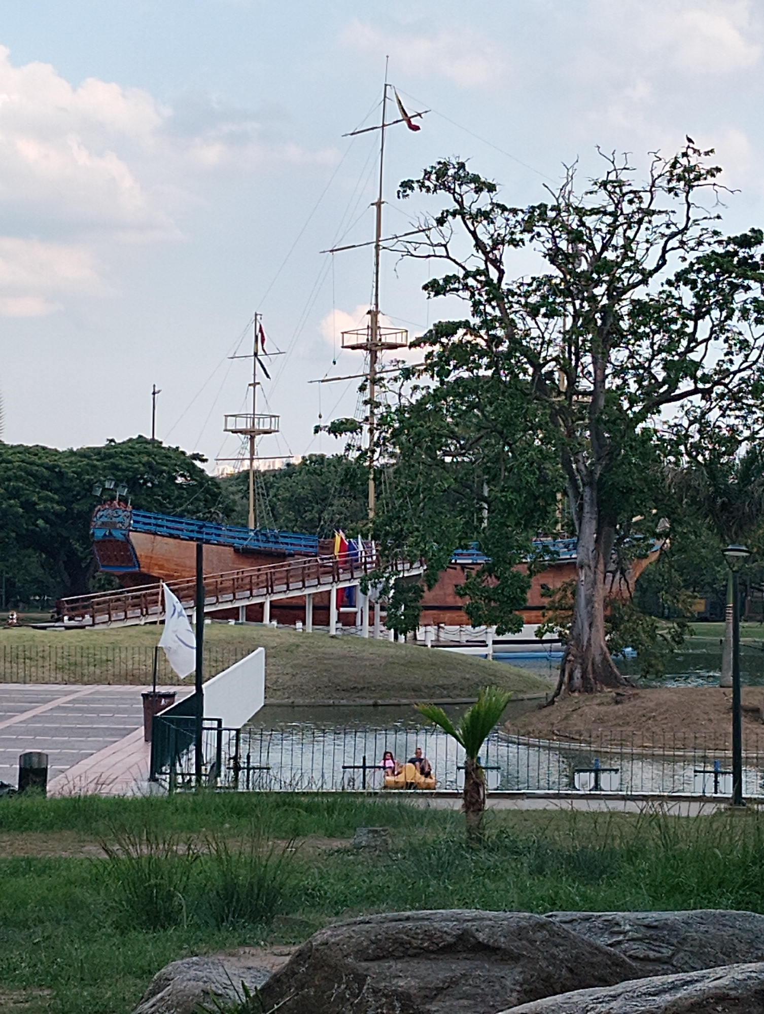 The image shows a static display of a large wooden galleon at Parque del Este in Caracas, Venezuela. The ship is positioned on an elevated mound and accessed by a wooden ramp. It has multiple masts and flags. A large tree stands to the right of the galleon, with bare branches at the top.
In the foreground, a small body of water is visible, with a metal fence separating it from the grassy area. Two people are seated in a yellow paddle boat in the water. Nearby is a small palm tree. A paved area with a white sign and a trash can are visible. Rocks are in the extreme foreground. The sky is cloudy.