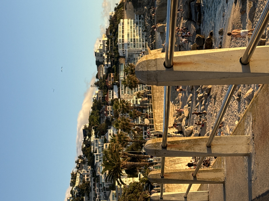The image captures a coastal scene in Cape Town, South Africa, likely within the Clifton or Camps Bay area, illuminated by warm, golden-hour sunlight. In the foreground, a weathered concrete promenade barrier with multiple posts and two horizontal stainless steel rails angles across the frame. Beyond this barrier, a sandy beach is interspersed with numerous large, grey rocks. Approximately 30-40 individuals are distributed across the sand and rocks, engaged in various leisure activities such as sitting, standing, and walking. One shirtless male in white shorts stands near the water's edge on the far right. The ocean is visible to the right, exhibiting small waves. In the midground, an elevated concrete retaining wall with additional people walking along its length separates the beach from a steeply rising hillside. The hillside is densely covered with multi-story residential buildings and modern houses, interspersed with lush green vegetation and numerous palm trees. A low-lying cloud formation is present along the upper slopes of the hill. The sky is clear blue in the upper portion, with several small birds visible in flight.