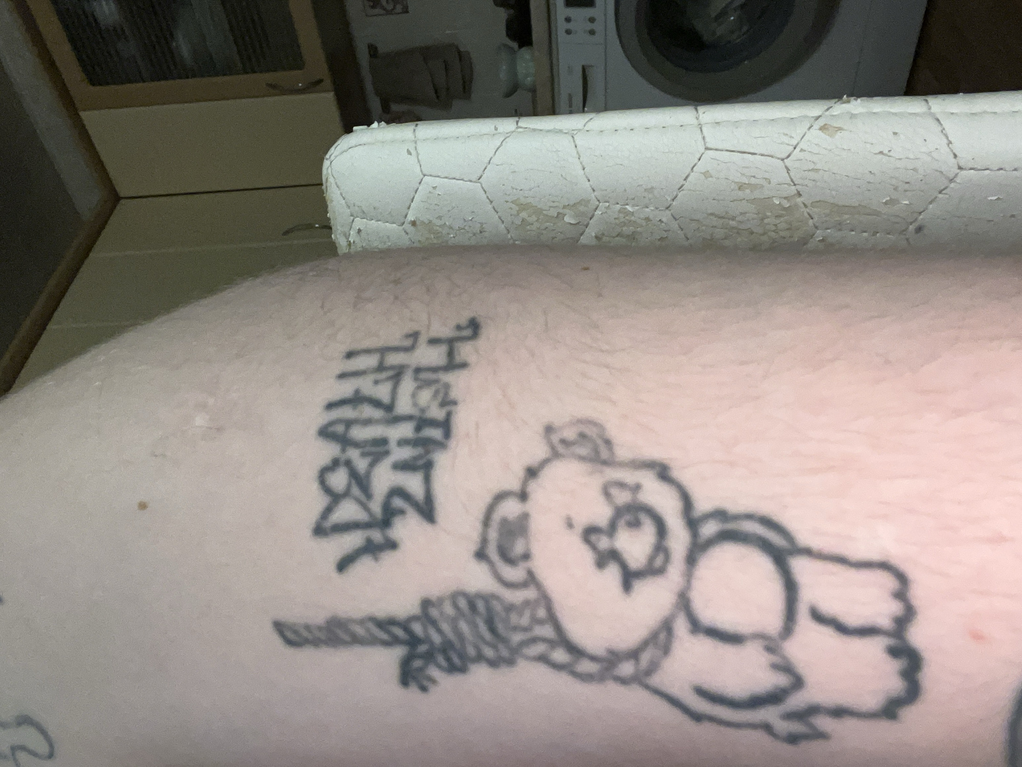 A tattoo of a teddy bear hanging from a noose is visible on a person's skin. Above the teddy bear tattoo, there is graffiti that reads "DEATH 2NITE". The person's skin is light-skinned and appears to have some texture. In the background, a washing machine and kitchen cabinets are visible. The overall environment suggests an indoor setting, possibly a home. The location is identified as Unknown, Unknown.