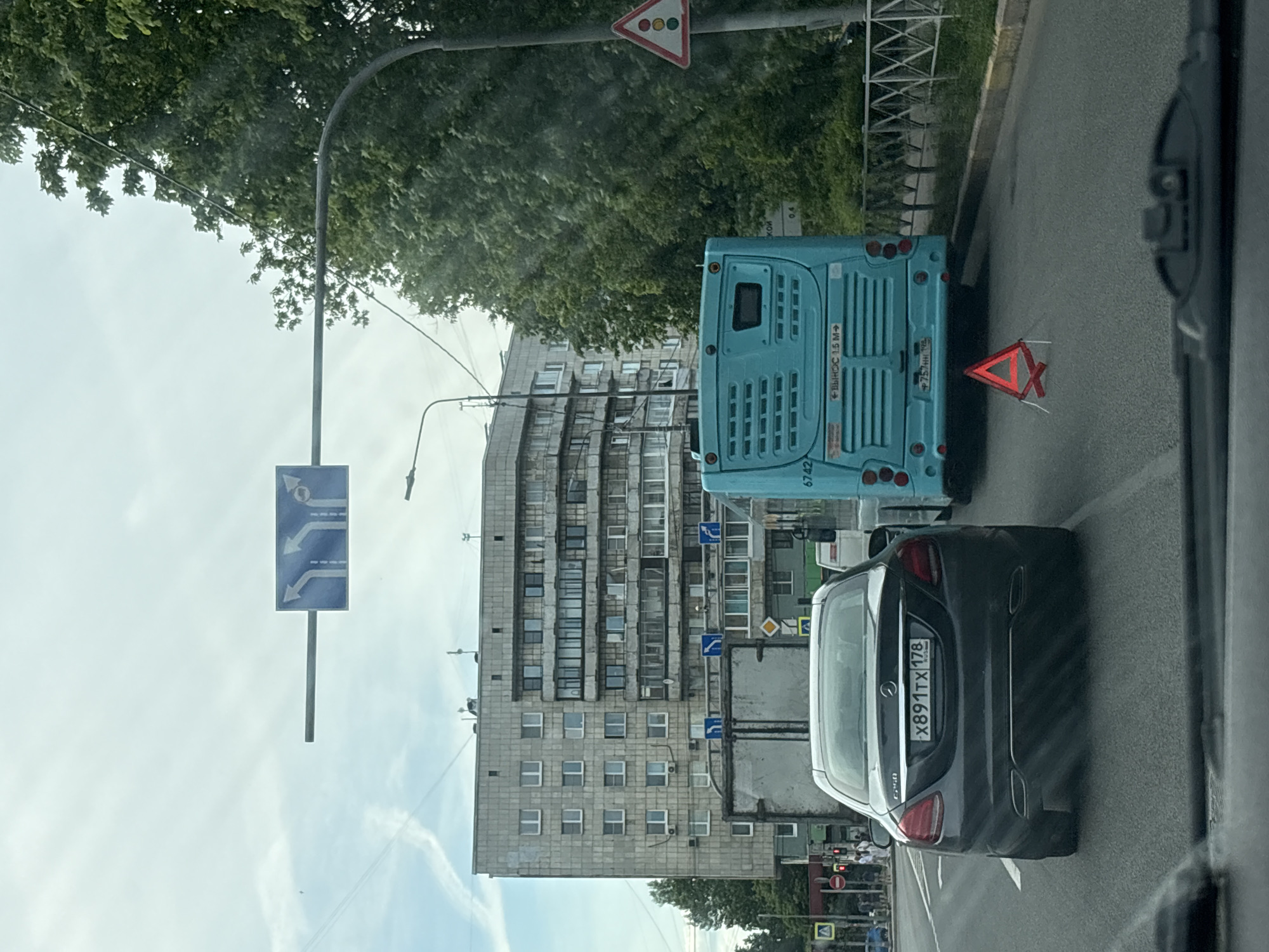 This daytime photograph captures an urban street scene in Saint Petersburg, Russia, observed from the interior of a vehicle looking forward. The sky is overcast and bright, with faint cloud streaks.
In the foreground, the top edge of a vehicle dashboard is visible. Immediately ahead, a dark grey Mercedes-Benz C-Class sedan (W205 generation) occupies the leftmost of three visible lanes. Its rear license plate, "X891TX 178 RUS," identifies its registration region as Saint Petersburg.
To the right of the Mercedes, a light blue urban bus, possibly a Volgabus model, is stopped or parked partially on the shoulder of the road. A red reflective emergency triangle stands on the asphalt directly behind the bus. The bus has the number "6742" on its rear side, and a warning sign on its rear window states "ВЫНОС 1.5 M→" (1.5 meter overhang). A partially visible rear license plate on the bus begins with "757" and ends with "198". Dense green trees line the right side of the road, behind the bus and a metal fence.
In the mid-ground, an overhead blue traffic sign with three white arrows indicates lane directions: the two left lanes proceed straight, while the rightmost lane turns right. Further down the street on the right, a triangular warning sign with three colored circles (red, yellow, green) indicates an upcoming traffic light.
The background features a large, multi-story (approximately 8-9 stories) grey residential building with a mix of older and newer windows and some enclosed balconies, indicative of Soviet-era architecture. Several smaller blue circular signs with white arrows (indicating mandatory turn directions) are visible near the base of this building. A yellow diamond-shaped sign (priority road) is also discernible. On the far left, a sidewalk with several pedestrians is visible, along with a blue "A" sign (pedestrian crossing) and a red circle "no entry" sign.
