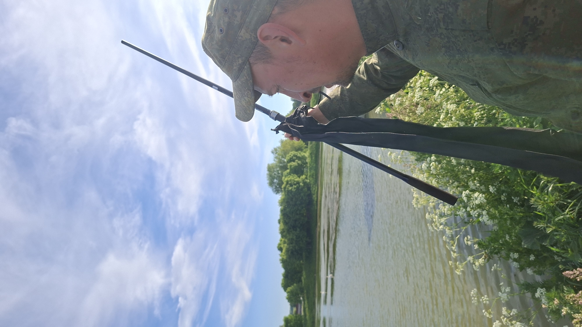A man in a camouflage cap and shirt is intensely focused on adjusting or setting up a fishing rod beside a body of water. He holds the rod with both hands, his gaze directed downwards towards the reel area. The fishing rod extends diagonally across the frame.
The setting is outdoors, by what appears to be a lake or river, with the water occupying the lower half of the original (rotated) image. The far bank across the water is lined with lush green trees and foliage, suggesting a natural and verdant environment. On the near bank, where the man is positioned, white wildflowers or small bushes are abundant, adding to the natural scenery.
The upper half of the original (rotated) image features a bright blue sky with scattered white, wispy clouds, indicating clear and sunny weather. The lighting suggests it is daytime, likely morning or afternoon. The man's posture and concentration imply he is engaged in fishing or preparing for it. There is no visible text in the image. The location context provided is Забалоцкі сельскі Савет, Belarus.
