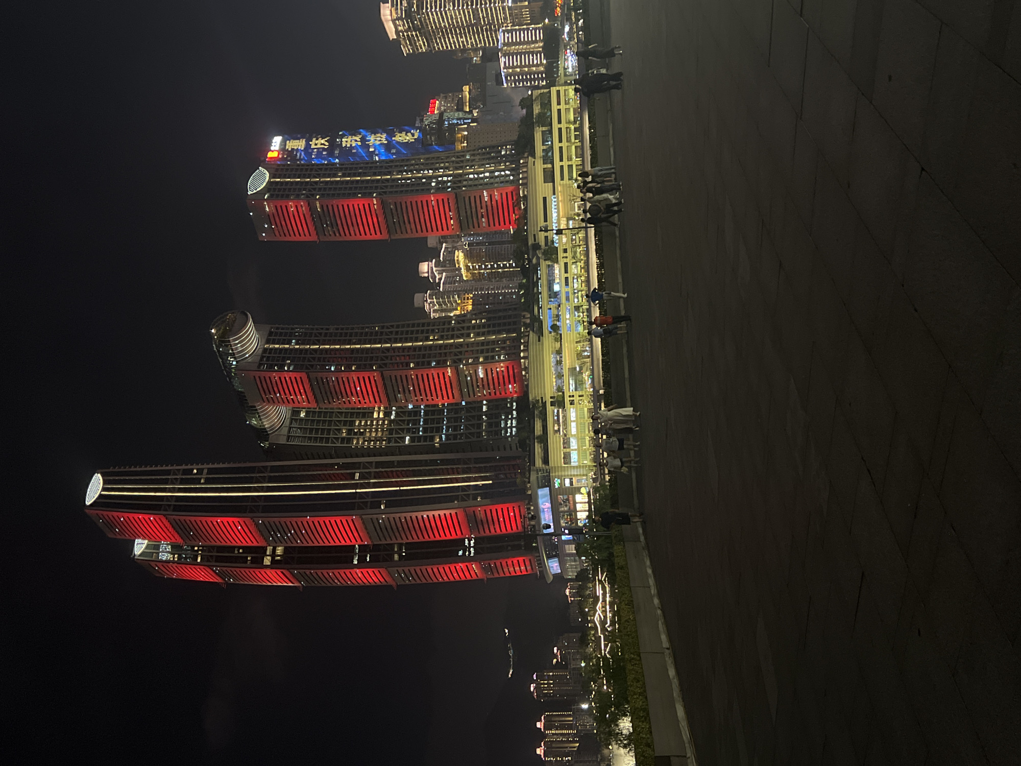 The image captures a nighttime view of a modern city skyline, featuring several illuminated skyscrapers. In the foreground, a dark paved area, possibly a plaza or walkway, is visible. A group of approximately ten people are scattered across this area, appearing to be casually walking or standing. Beyond the paved area, a lower building with extensive lighting is present, with a line of green foliage separating it from the foreground. In the background, a series of skyscrapers dominate the scene, with some featuring striking red and blue lighting. One of the taller buildings displays Chinese characters in blue light. Further in the distance, other buildings are lit up, contributing to a vibrant urban nightscape. An airplane is faintly visible in the sky to the left of the frame. The overall impression is a bustling city at night with contemporary architecture. The location is identified as Jiangbeicheng Street, China.