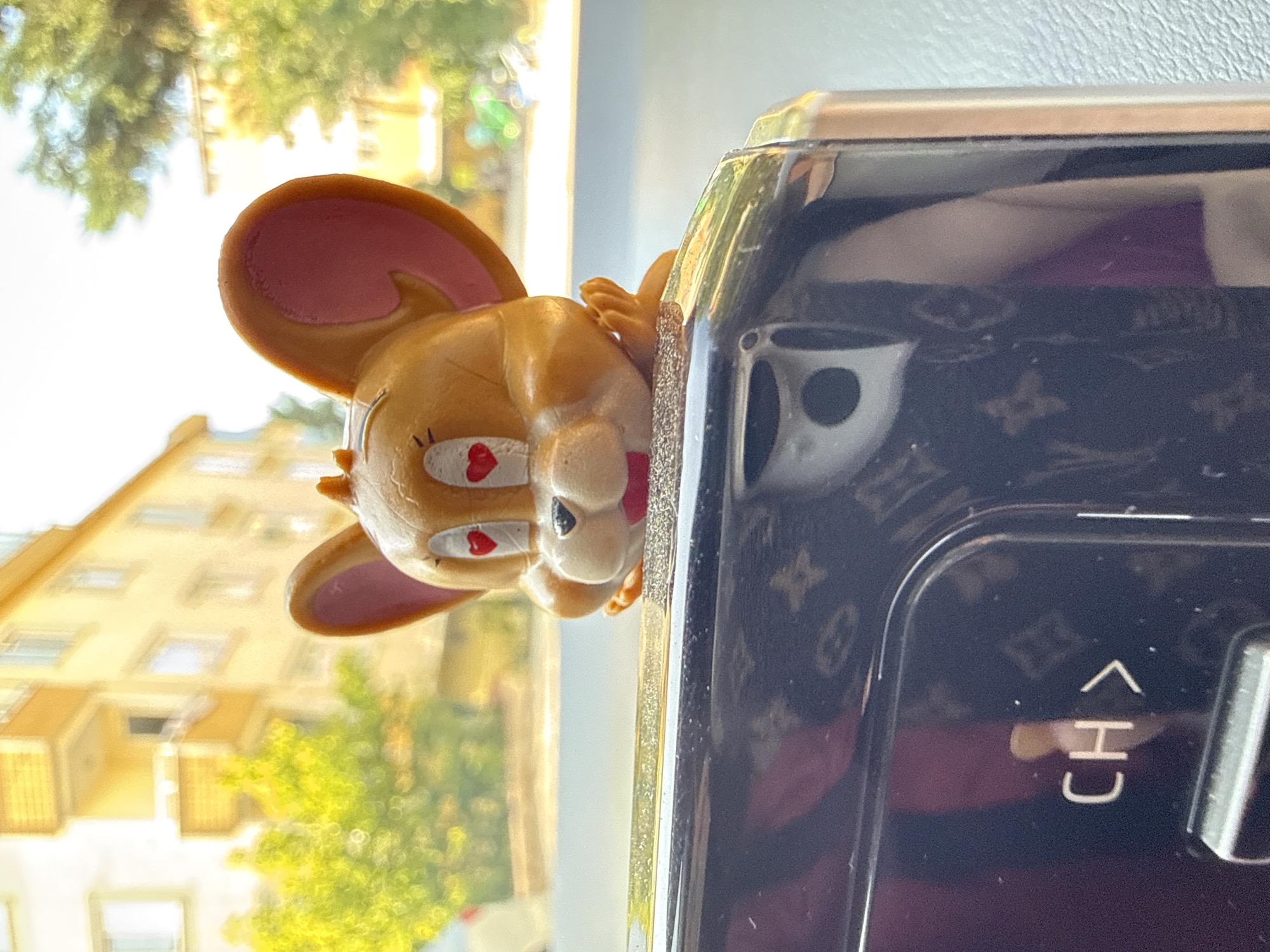 An outdoor scene in Sancaktepe, Turkey, features a close-up shot of a Jerry figurine with heart-shaped eyes, placed on the dashboard of a vehicle. The figurine is positioned above a dark, reflective car dashboard with a repeating pattern. In the background, a building with yellow facades and trees with green foliage are visible, suggesting an urban environment. The lighting indicates it is daytime.