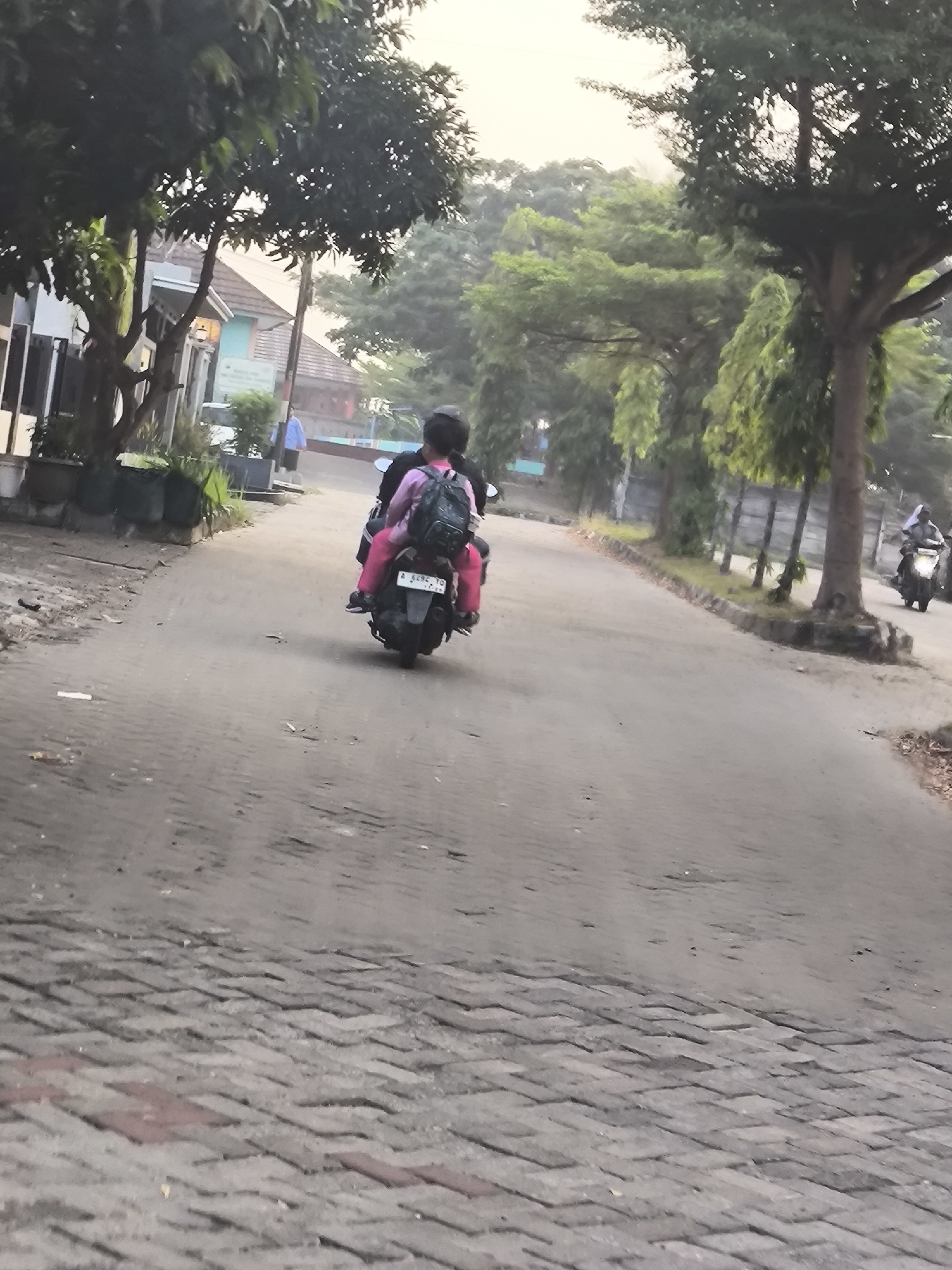 The image depicts a street scene in Cilegon, Indonesia. In the foreground, a paved road transitions to a cobblestone surface. Two people are riding on a motorcycle, with the rider in the back wearing a pink shirt and black backpack. The motorcycle has a license plate that appears to read "A 6292 TO". On the left side of the road, there are houses and trees. On the right side, there are more trees and a fence. In the distance, another motorcycle is visible. The sky is overcast, suggesting it might be early morning or late afternoon.