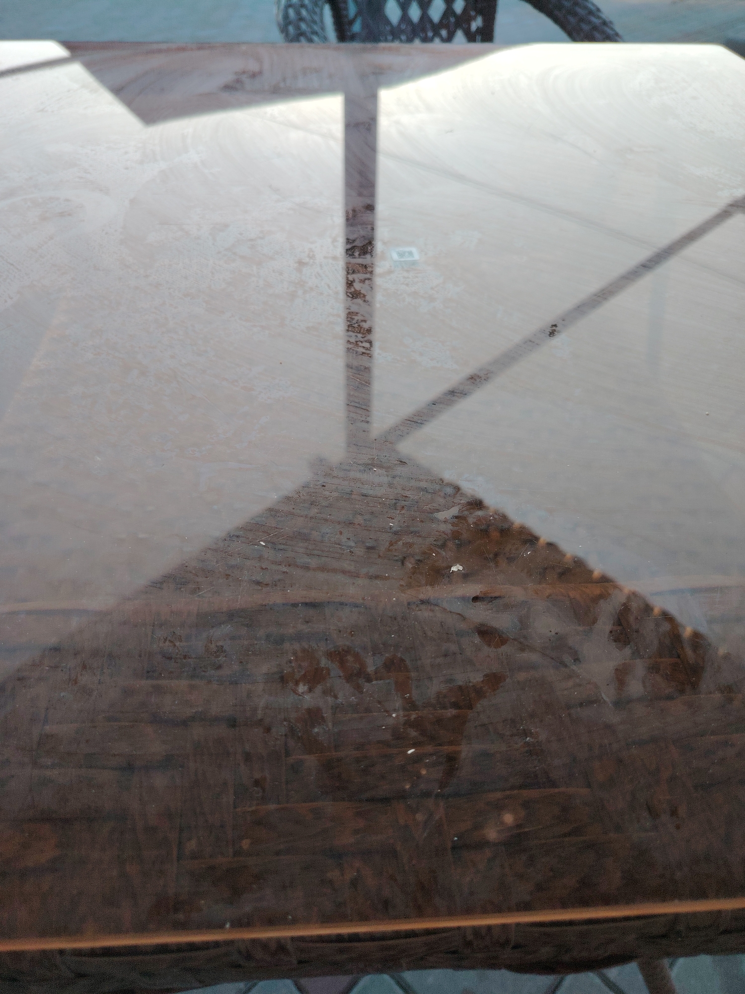The image shows a close-up view of a wooden table with a glass top. Sunlight creates distinct shadows on the table's surface, indicating an outdoor setting. The wooden surface beneath the glass appears to be made of planks, with a weathered or textured finish. Some small debris or marks are visible on the table. In the background, out of focus, the edge of a tire or wheel and a portion of a wicker-like structure are visible, suggesting the presence of furniture or a vehicle nearby. The overall scene is likely a patio or outdoor seating area. The location is identified as Toranghyliq, Kazakhstan.