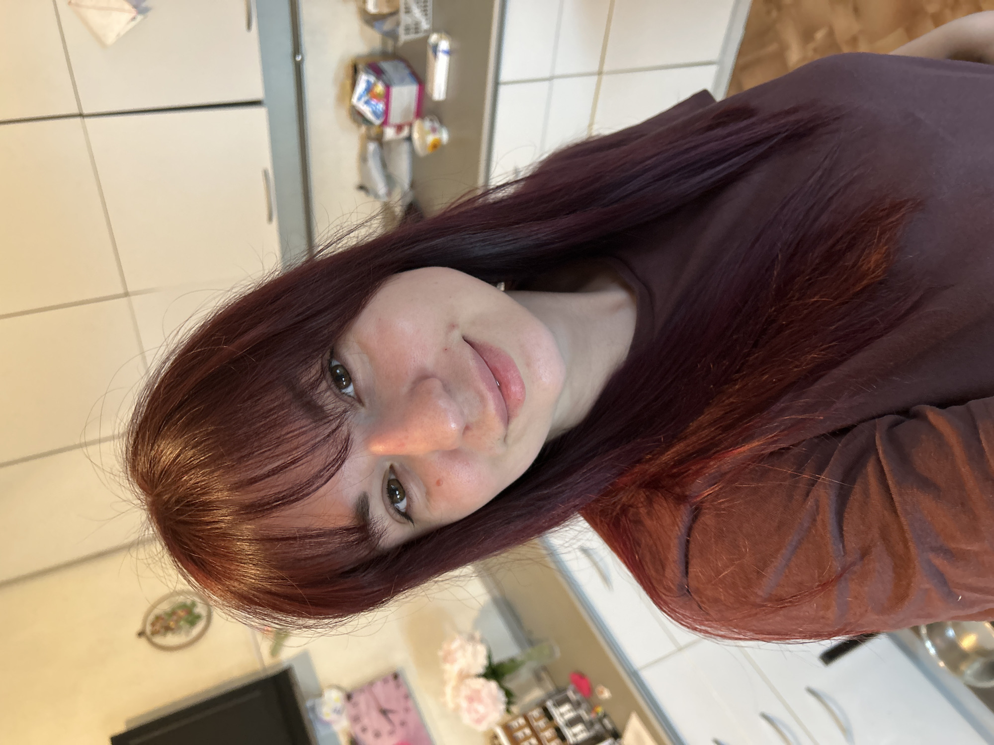 A woman with long, auburn hair and bangs is taking a selfie in a kitchen. She is wearing a dark maroon t-shirt. The background shows white kitchen cabinets, a countertop with various items, and a framed piece of art on the wall. There are also some flowers in a vase and a clock on the wall. The image does not provide any information about the city of Фоки, Russia.