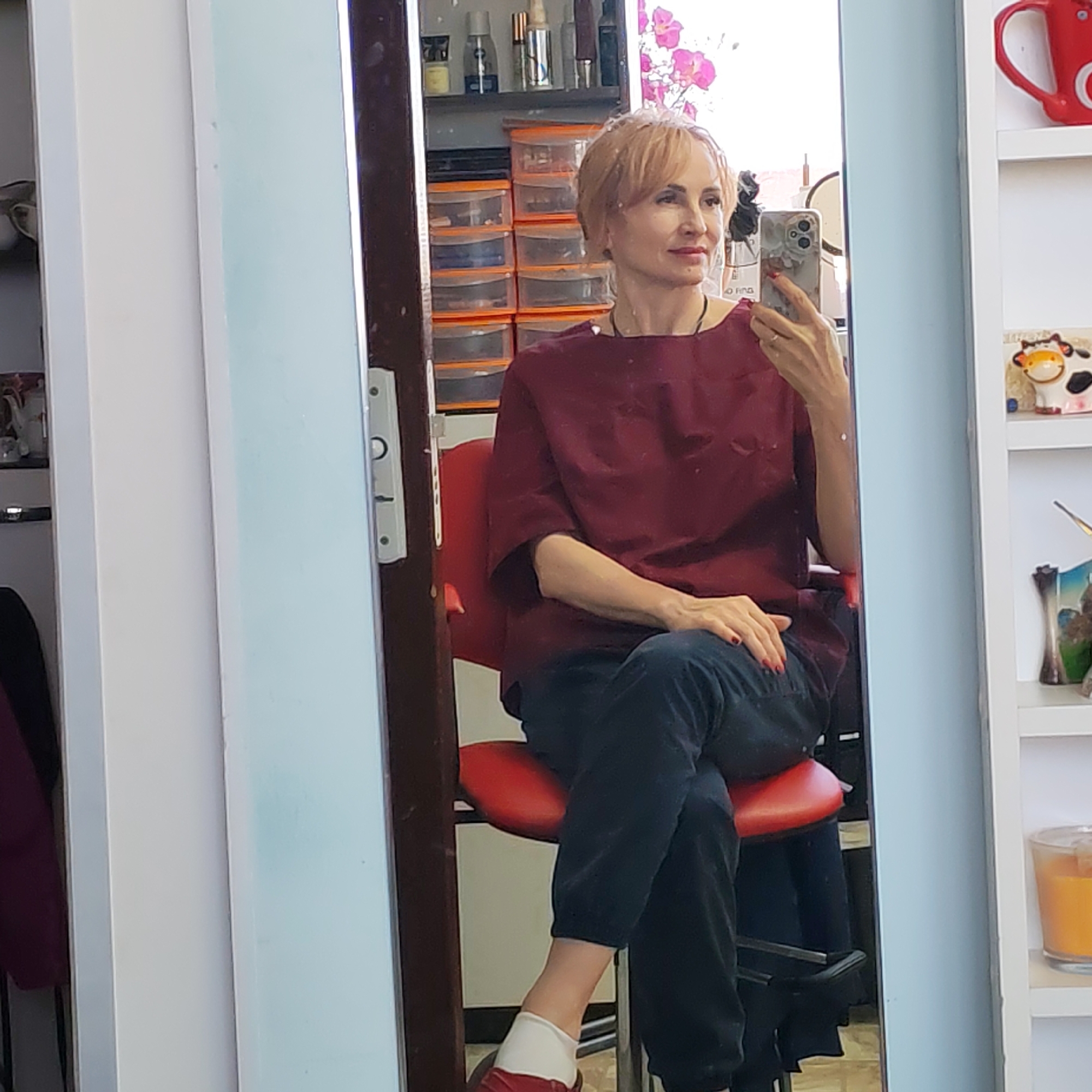 A woman with short, light-colored hair is sitting on a red, high-backed chair in a room that appears to be a salon or possibly a bathroom. She is wearing a dark red, loose-fitting shirt and dark-colored pants rolled up to the ankle, paired with white socks and red shoes. She is holding a smartphone in her right hand, taking a selfie in a large mirror in front of her. The mirror reflects a cluttered shelf of beauty products and personal care items, as well as orange storage drawers. A second mirror is visible to the left. To the right of the mirror is a white shelving unit containing a red mug with a "C" logo and a ceramic cow figurine. The door hardware of the mirror indicates that it is located in Russia. The location is Chaikovsky, Russia.