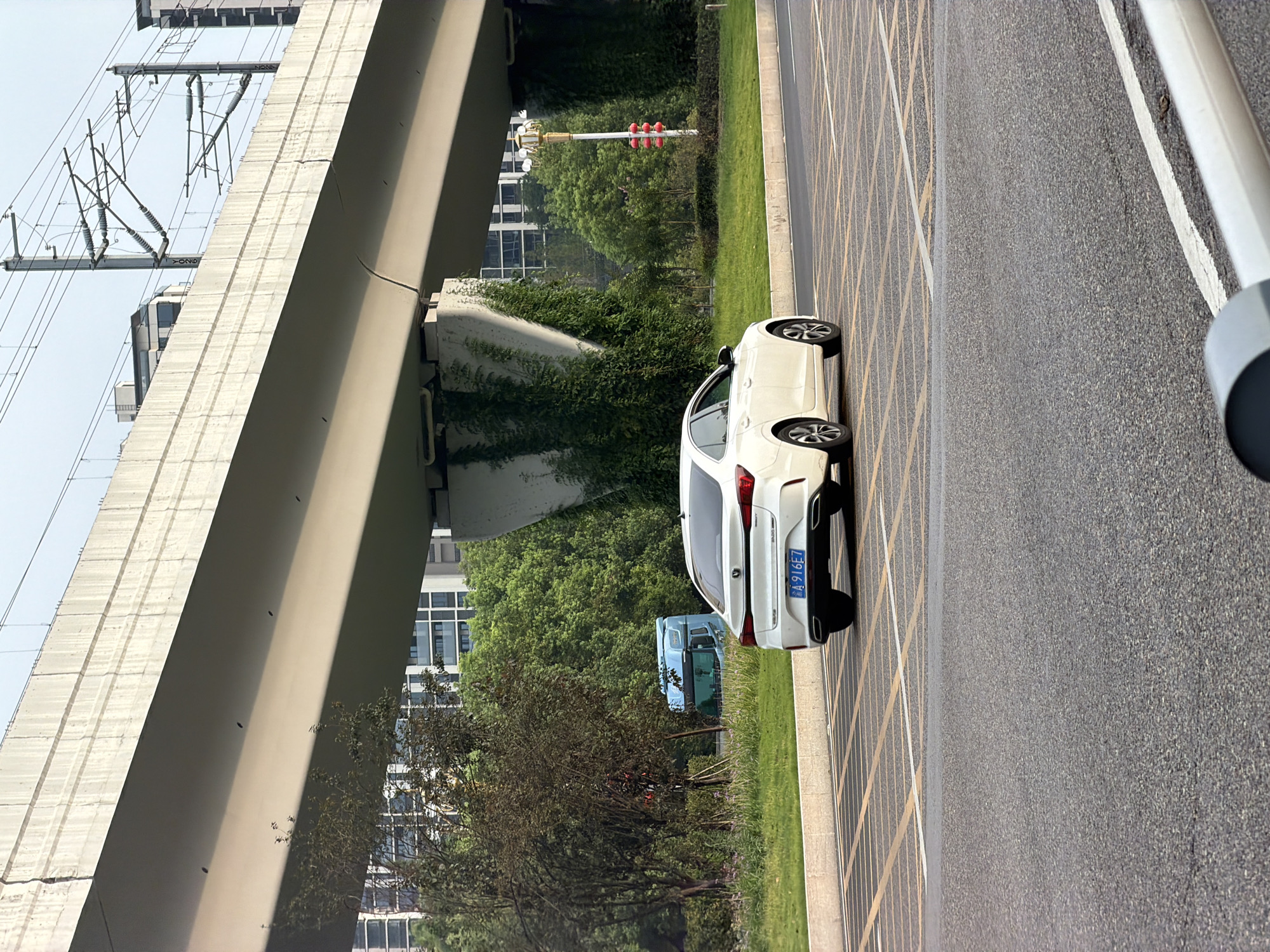 In the city of Jiangjin, China, a white sedan with license plate "A 916E7" is parked in a marked parking area. The car is viewed from the rear, angled slightly towards the right. Behind the car, a light blue truck is visible, partially obscured by trees. A raised concrete overpass dominates the upper portion of the image, with overhead power lines for an electric train. The environment appears to be a roadside with green grass and trees. The sky is overcast.