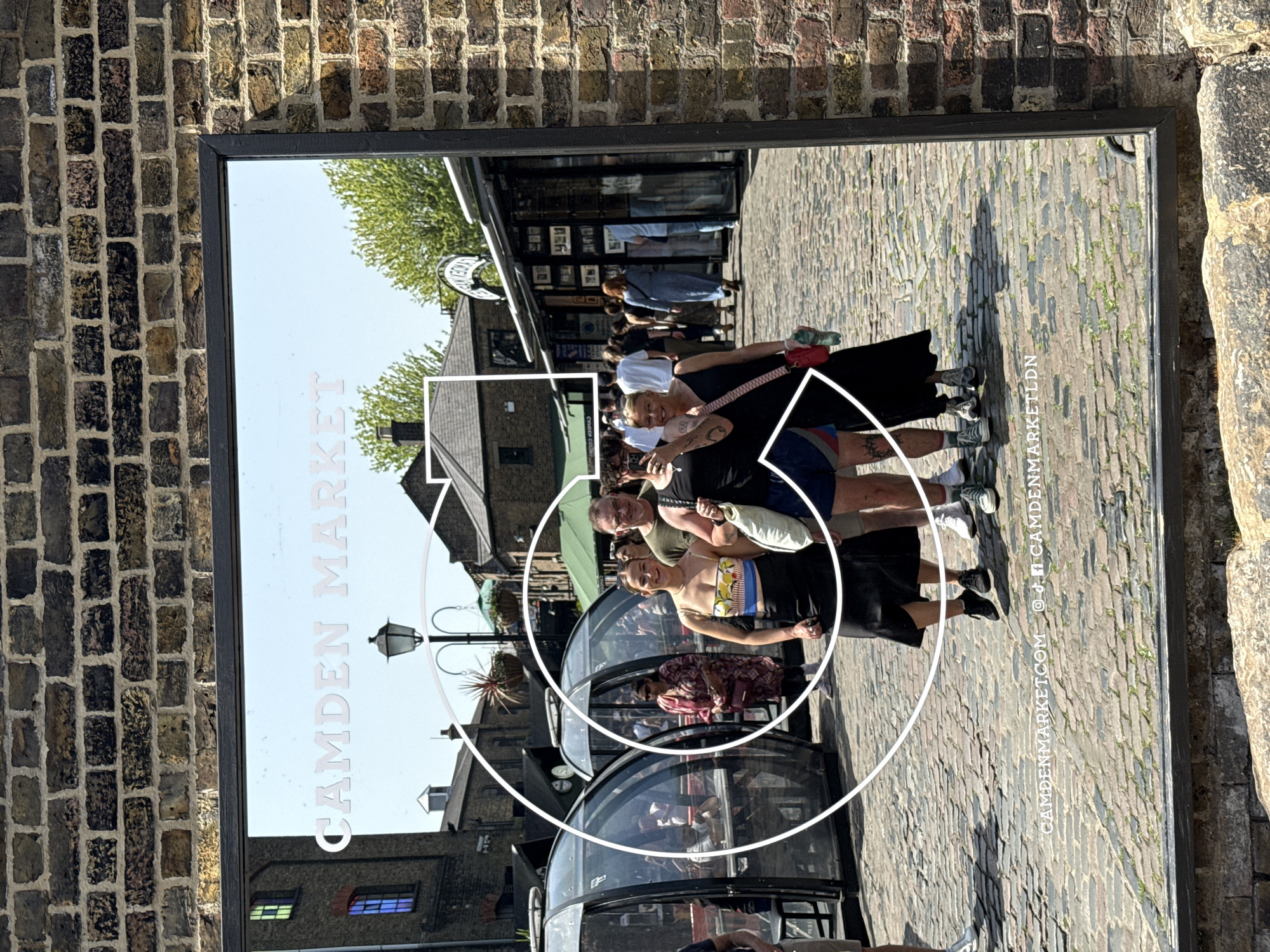 A framed promotional poster for Camden Market in Camden Town, London, is affixed to a brick wall. The poster features a photo of a group of people standing in front of the market's distinctive cylindrical shops on a cobblestone street, appearing to pose for the shot. The words "CAMDEN MARKET" are printed vertically on the left side of the poster in white font, and a large, stylized graphic is overlaid on the image, partially obscuring the people. The poster also includes the market's website address and social media handles at the bottom right corner.