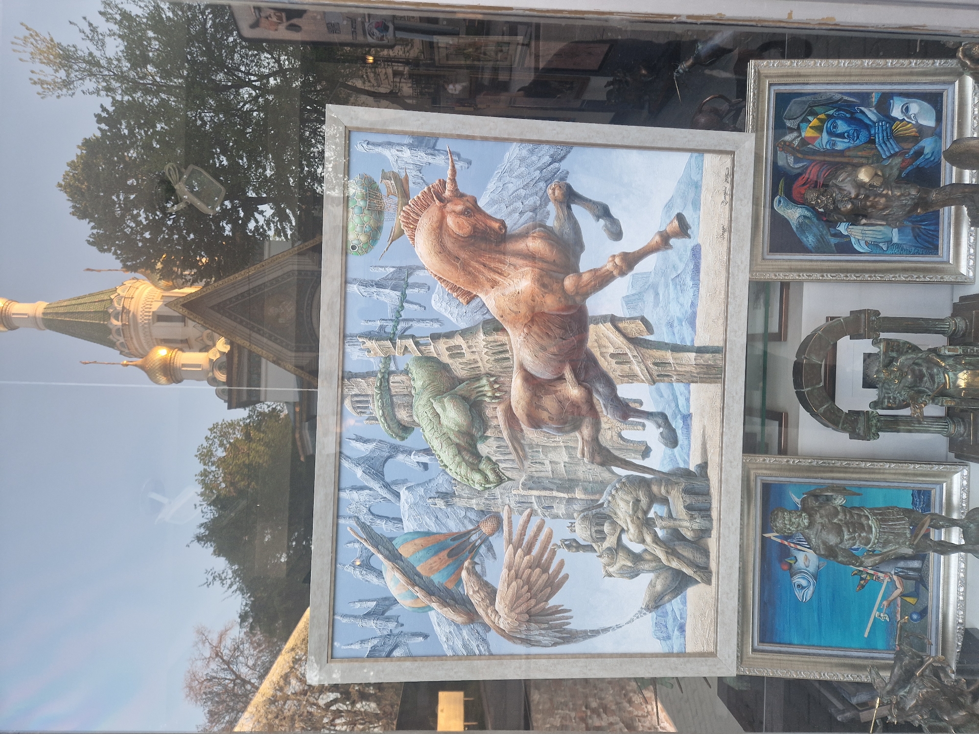 The image shows a street scene in Unknown, Unknown, viewed through a store window displaying artwork.
In the foreground, a large framed painting dominates the view. The painting features a surreal scene with a muscular, rearing unicorn-like creature, a serpentine dragon-like figure, winged orbs, and human-like figures in a fantastical landscape. The painting is in a silver-colored frame.
To the right of the large painting, other framed artworks are visible. These include depictions of human figures in various poses and contexts. Bronze sculptures are partially visible in front of and to the side of these paintings.
The window's reflection captures elements of the street. A building with a gold-domed spire is reflected, as are trees with sparse foliage, possibly indicating autumn or winter. A partial reflection of the sky is visible in the upper portion of the window.