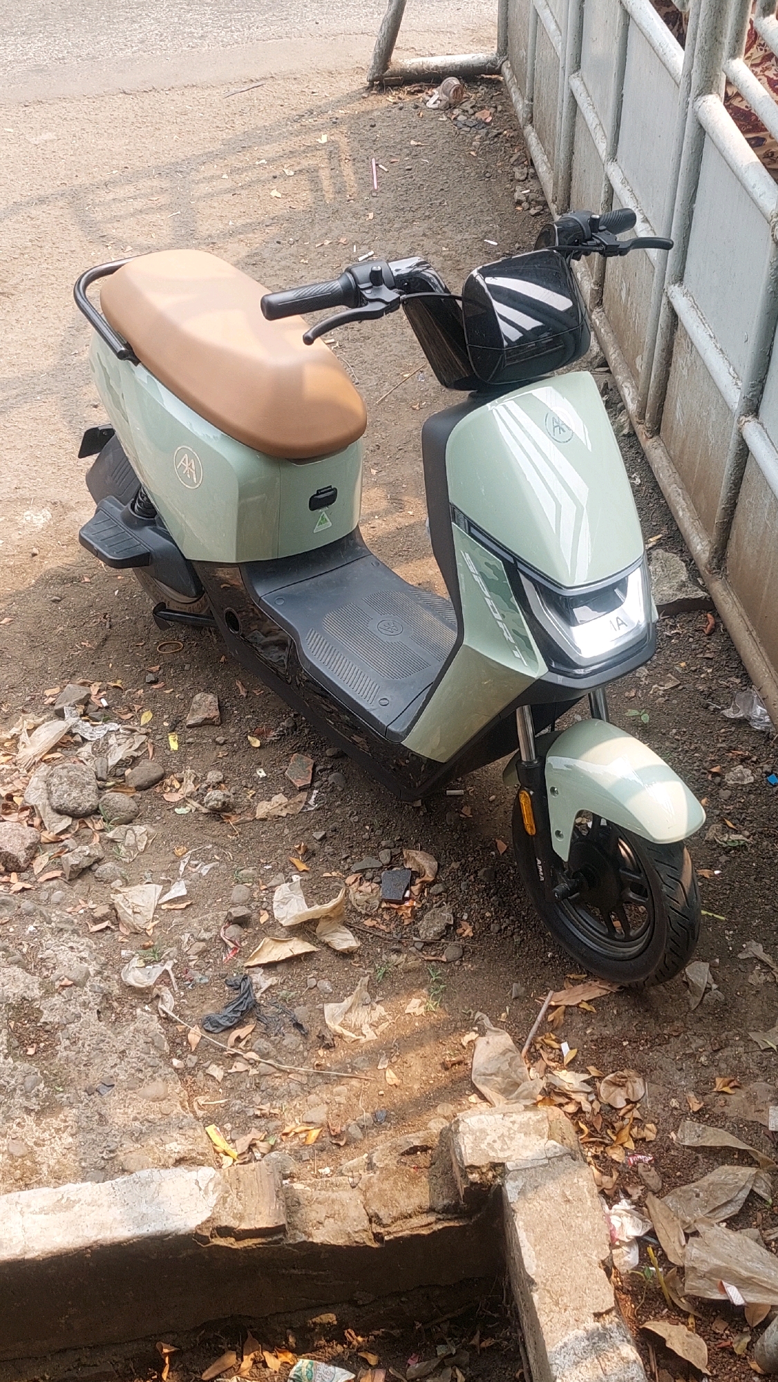 An electric scooter is parked on a dirt road in Babakan Madang, Indonesia, with a light green body and a tan seat. The scooter has a black handlebar with grips and levers, and a black front tire. The scooter is positioned to the left of a light gray metal fence. The ground around the scooter is littered with debris, including leaves, plastic bags, and small stones. In the foreground, there is a concrete structure with a rectangular opening. The background shows a paved road and more debris. The lighting suggests it is daytime.