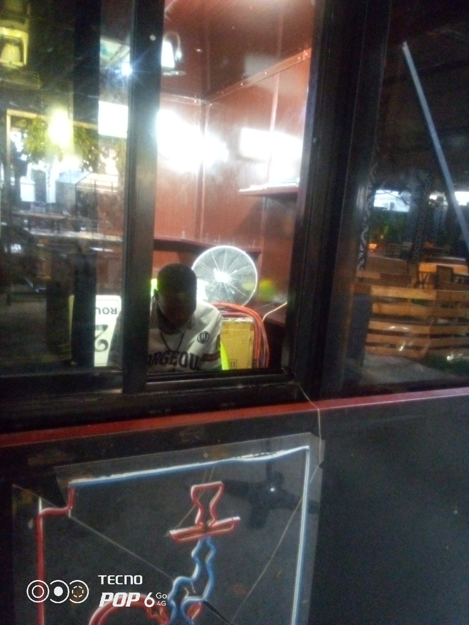 The image captures a dimly lit indoor scene, likely a business establishment in Howayi, Nigeria, viewed through a window. In the foreground, a portion of a glass enclosure with red trim is visible, reflecting light and displaying some sort of neon signage or decoration with red and blue elements at the bottom. A watermark indicates "TECNO POP 6 4G".
Behind the glass, a person is seated, their face obscured by shadow and their head bowed. They appear to be wearing a light-colored shirt with dark lettering that reads "RGEOU". A white sign with black lettering, partially visible and rotated, shows "ROU" and "G". To the right of the person, a white electric fan is operating, and behind it are yellow boxes and some coiled orange and red wires. The background through the window suggests an outdoor area with tables and chairs, possibly a seating area for patrons, and what appears to be wooden pallet furniture. The overall impression is of an establishment operating at night or in low light conditions.