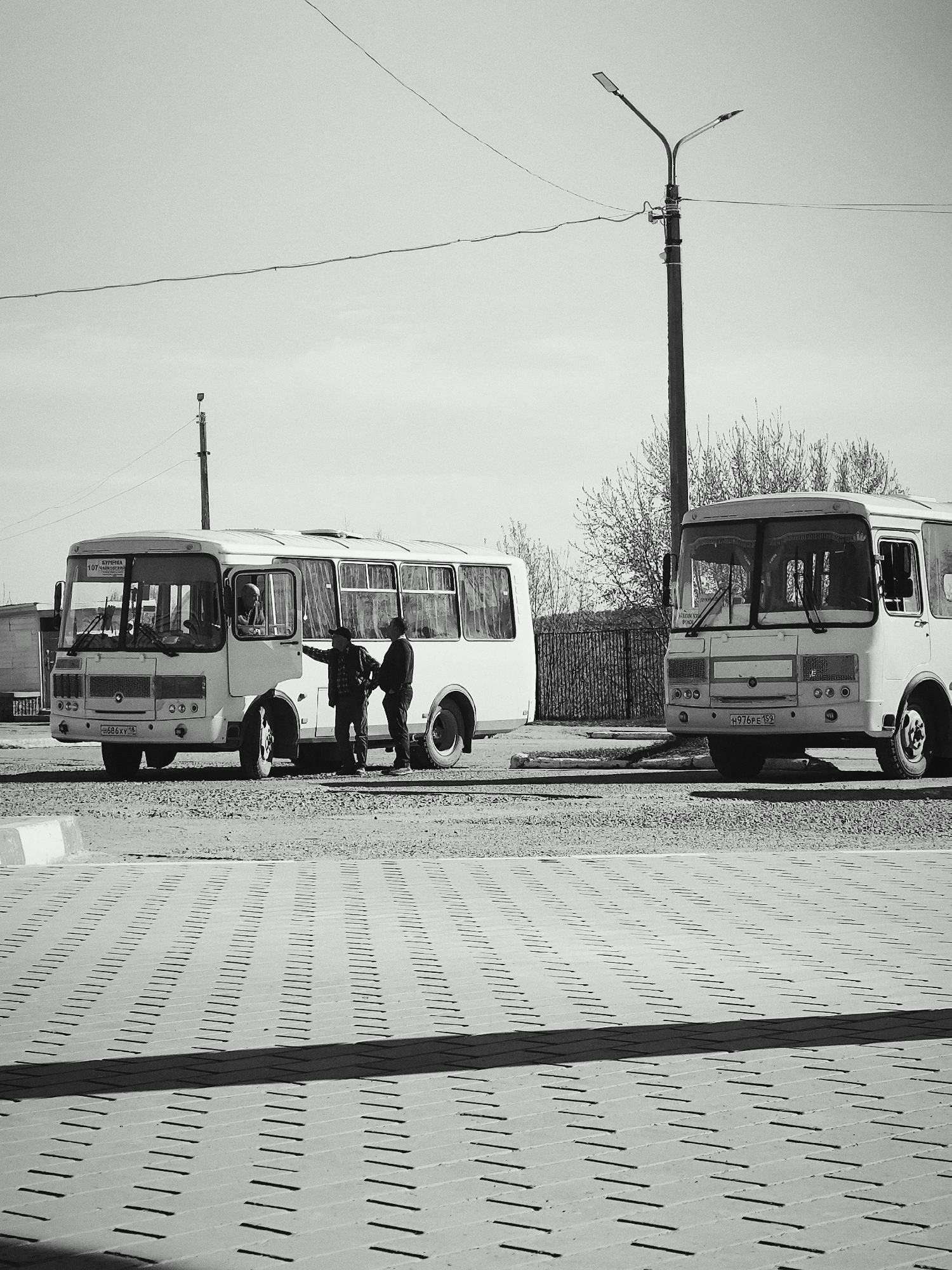The black and white image shows two white buses parked on a paved area, likely a bus stop or terminal, in Chaikovsky, Russia. Two men stand beside one of the buses, appearing to be in conversation. A third person can be seen in the driver's seat of the bus. A street light is visible in the background, and a wire stretches across the sky. A fence and some trees can be seen behind the buses. The foreground is a paved area with a patterned brick design.