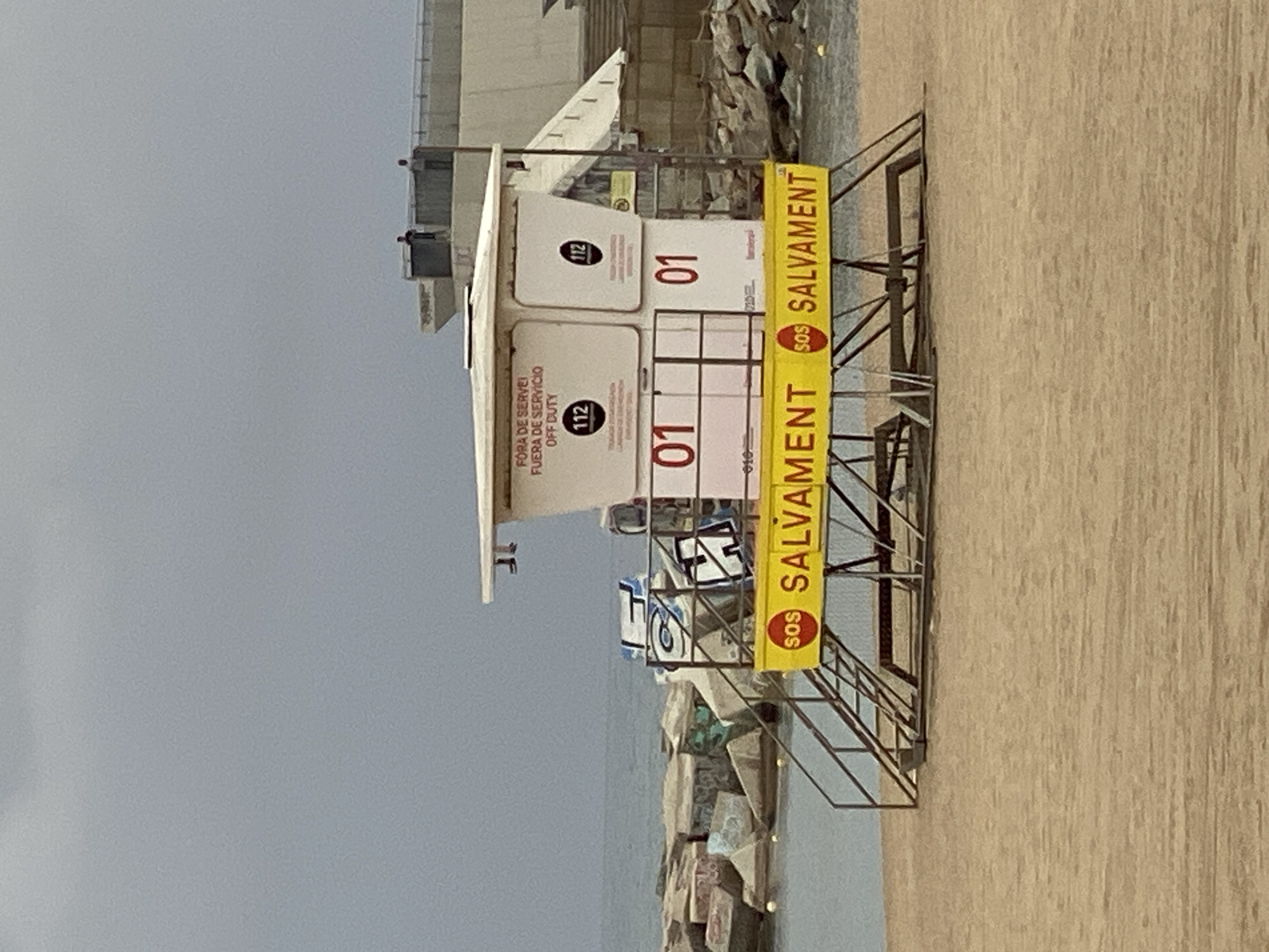 This media file captures a white, two-story lifeguard tower situated on a sandy beach. The tower features a metal staircase leading up to its main level. A prominent yellow banner, running along the front railing, displays "SOS SALVAMENT" in red and black lettering, with "SALVAMENT" being Catalan for 'rescue'.
On the white panels of the tower, multilingual text is visible, indicating its operational status: "FORA DE SERVEI" (Catalan), "FUERA DE SERVICIO" (Spanish), and "OFF DUTY" (English), all signifying that the station is currently out of service. The emergency number "112" is also displayed twice, along with the station identification number "01" appearing on two panels. Smaller text, "010", is faintly visible.
The scene depicts a coastline with a calm, grey-blue sea. A breakwater, constructed from large rocks, extends into the water to the right of the tower. In the background, a concrete structure or building can be seen. The sky is overcast and grey, suggesting a cloudy day. The sandy beach in the foreground appears undisturbed. No individuals are present in the image, contributing to a quiet, possibly off-season or early morning atmosphere. The visible text in Catalan and Spanish, combined with the context, suggests a location in Barcelona, Spain.