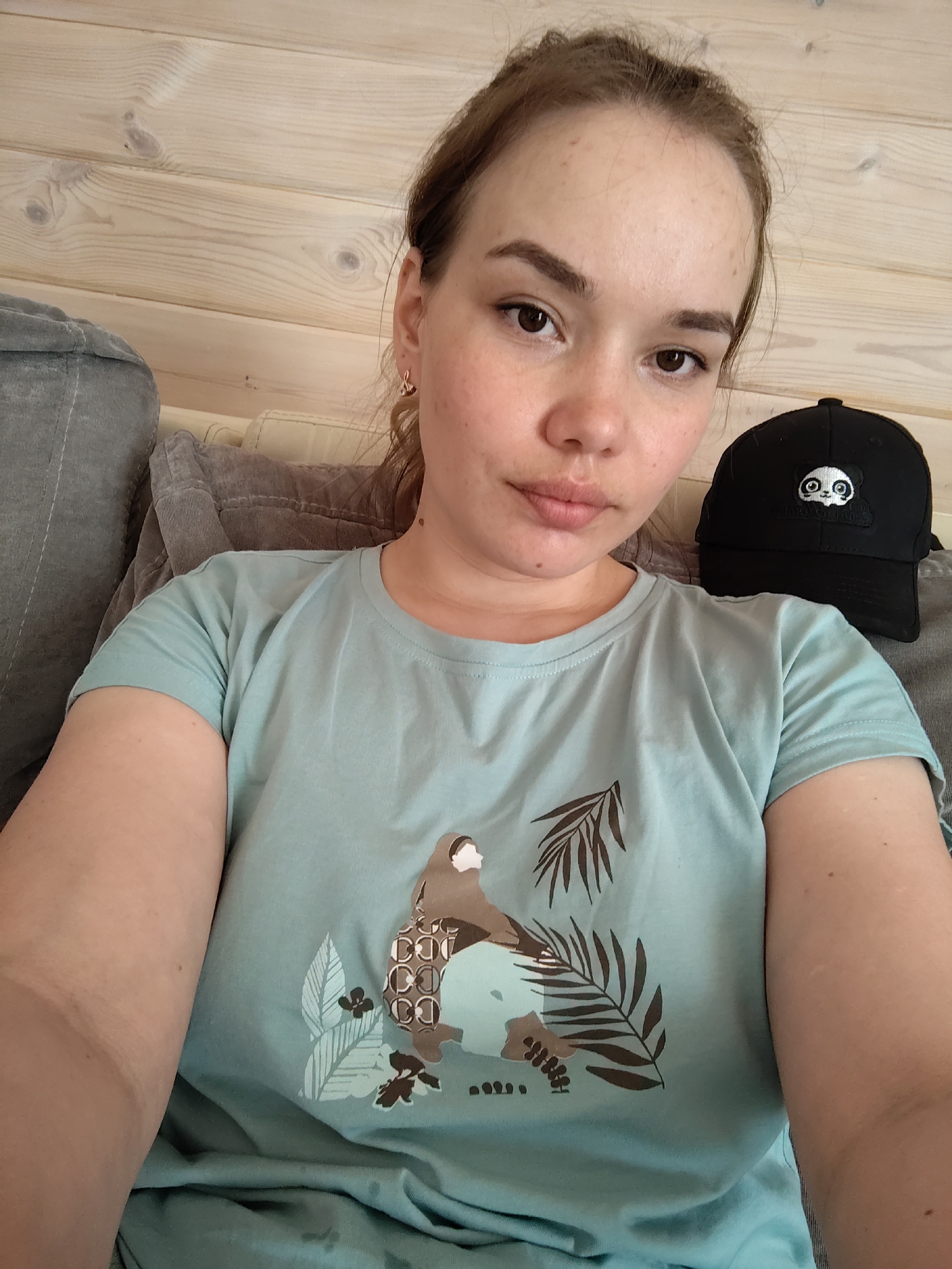 A woman with fair skin and light brown hair pulled back is centered in the frame, looking directly at the camera with a neutral expression. She wears a light blue t-shirt with a printed design of a bird and botanical elements. Behind her to the right is a black baseball cap with a panda bear design. She appears to be sitting on a gray upholstered sofa, with a light wood-paneled wall visible in the background. No definitive location within the city of Malyi Bukor, Russia can be determined.