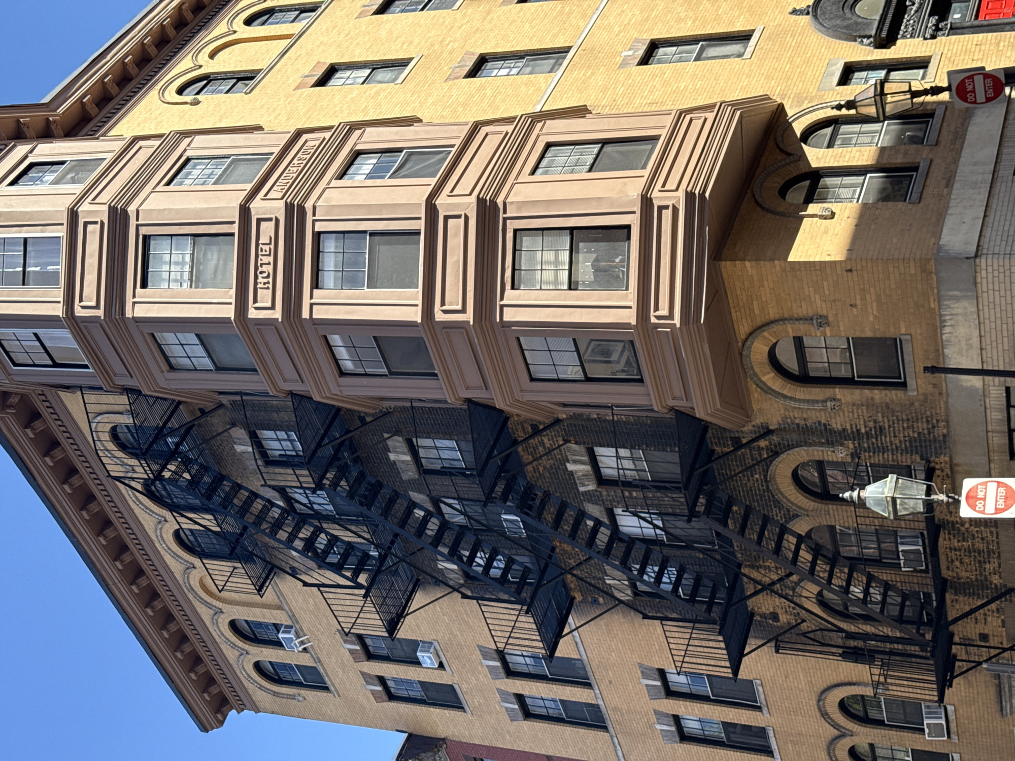 The image shows the Anderson Hotel building located in the city of Boston, United States of America. It features a yellow brick facade with a bay window section that has the inscription "ANDERSON HOTEL". A black fire escape ladder is affixed to the building's exterior. A "DO NOT ENTER" sign and a vintage-style streetlight are also visible near the building. The sky is blue and clear.