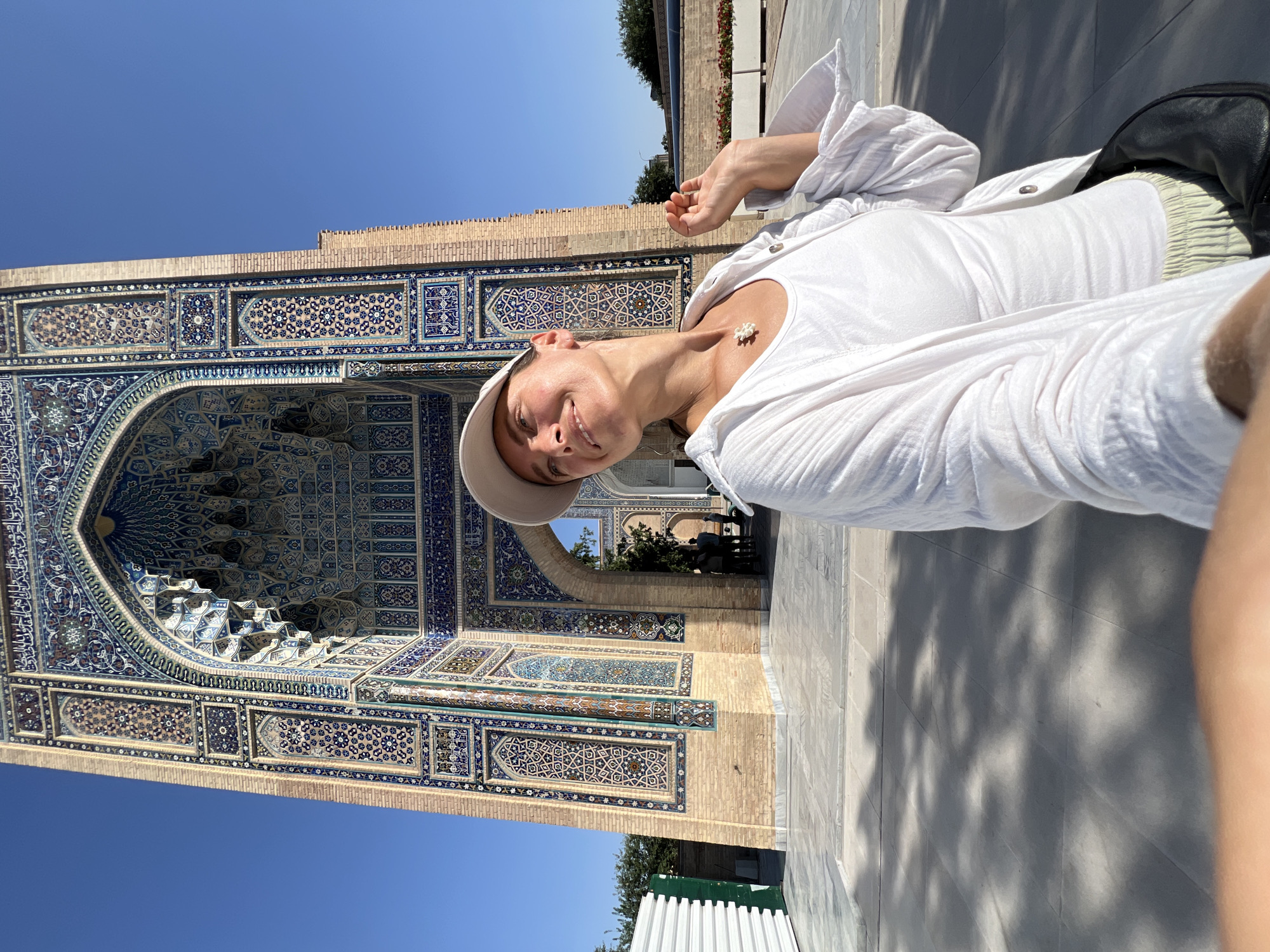 A person wearing a white shirt, a beige cap, and light-colored pants is taking a selfie in front of a building with intricate blue and white tilework. The building appears to be a historical or religious site, possibly a mosque or mausoleum, with ornate arches and geometric patterns adorning its facade. The sky is clear and blue, suggesting a sunny day, likely midday due to the strong shadows cast on the ground. The person is smiling and looking at the camera, appearing relaxed and happy. The setting appears to be an outdoor courtyard or plaza with paved ground. The tilework is a dominant feature, showcasing detailed Arabic calligraphy and geometric designs in shades of blue, white, and turquoise, against a backdrop of brick. In the background, glimpses of trees and other structures can be seen, hinting at a larger complex.