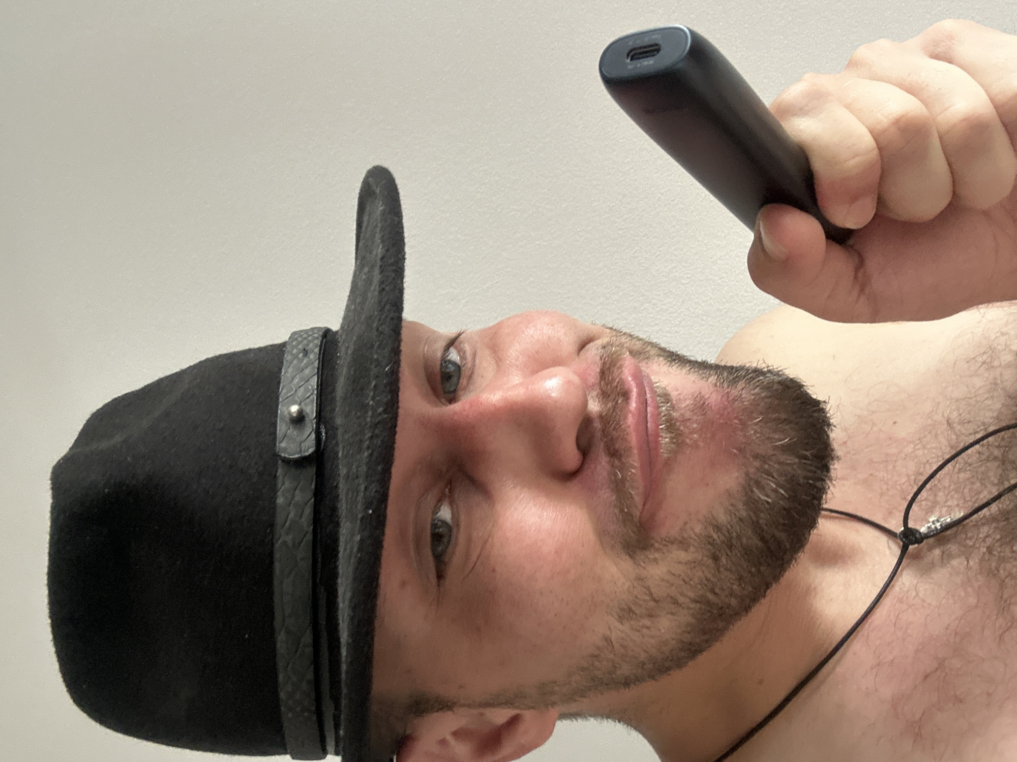 A man wearing a black fedora and a necklace with a cross pendant is holding a dark grey cylindrical object, possibly an electronic cigarette or a stylus, in his right hand. He has a beard and is looking towards the camera with a slight smile. The background is a plain, light-colored wall. The image appears to be a selfie, taken indoors. There is no discernible location within the city of Zvezdara Urban Municipality, Serbia.
