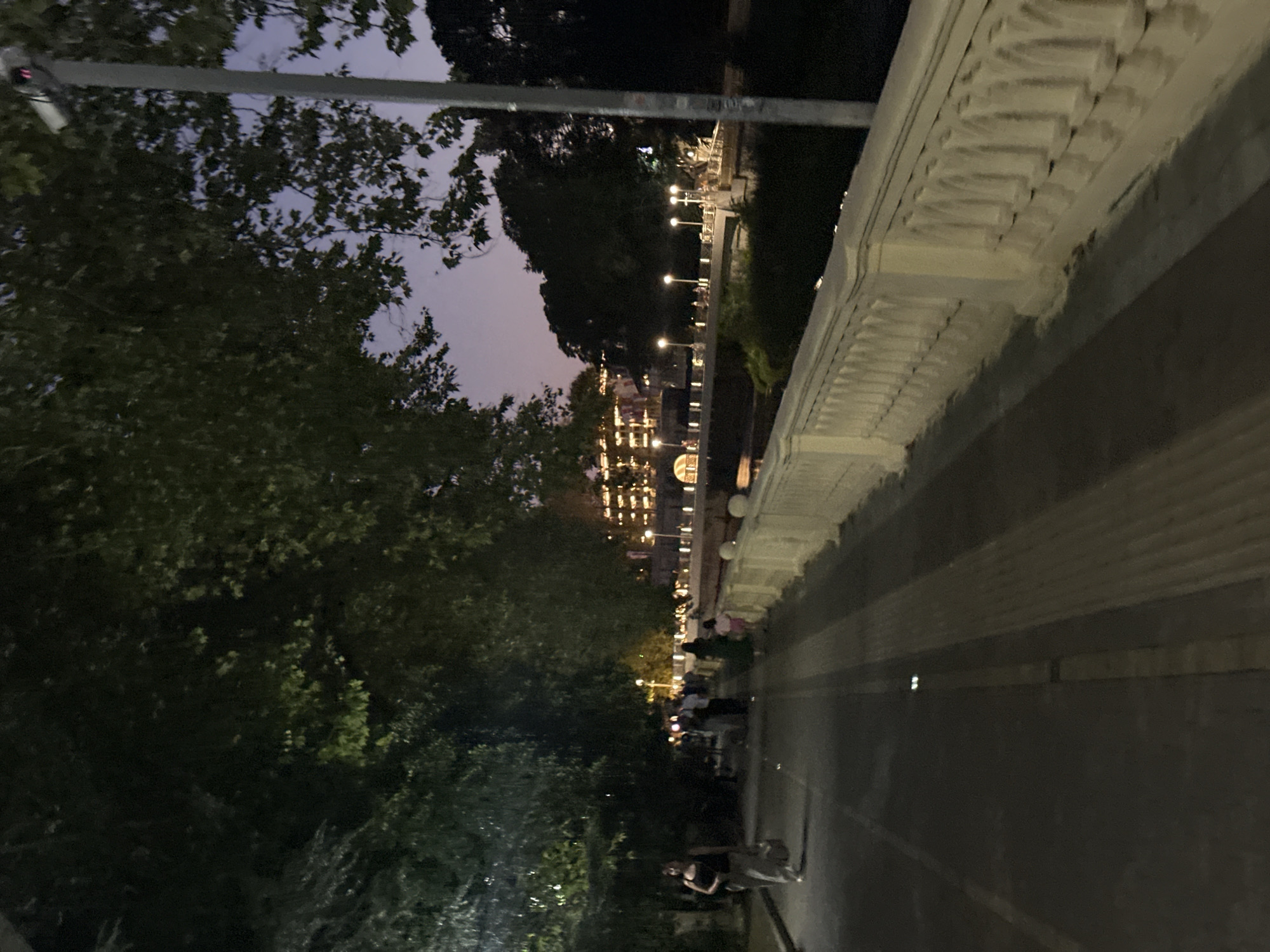 The image depicts a dimly lit pedestrian walkway next to a river or canal, possibly in Sochi, Russia, given the context provided. The time appears to be dusk or early evening, with the sky showing shades of purple and deep blue. Lush green trees with dense foliage overhang the walkway on the left, casting shadows. On the right, a decorative, light-colored railing lines the path along the water. Across the water, a bridge with illuminated buildings and streetlights is visible. Several people are walking along the path, some appearing to be tourists or locals enjoying an evening stroll. The walkway itself is paved with a dark surface, with lighter-colored sections and some embedded lights. A lamppost with a security camera attached is present on the right side of the image, adding to the urban infrastructure detail.