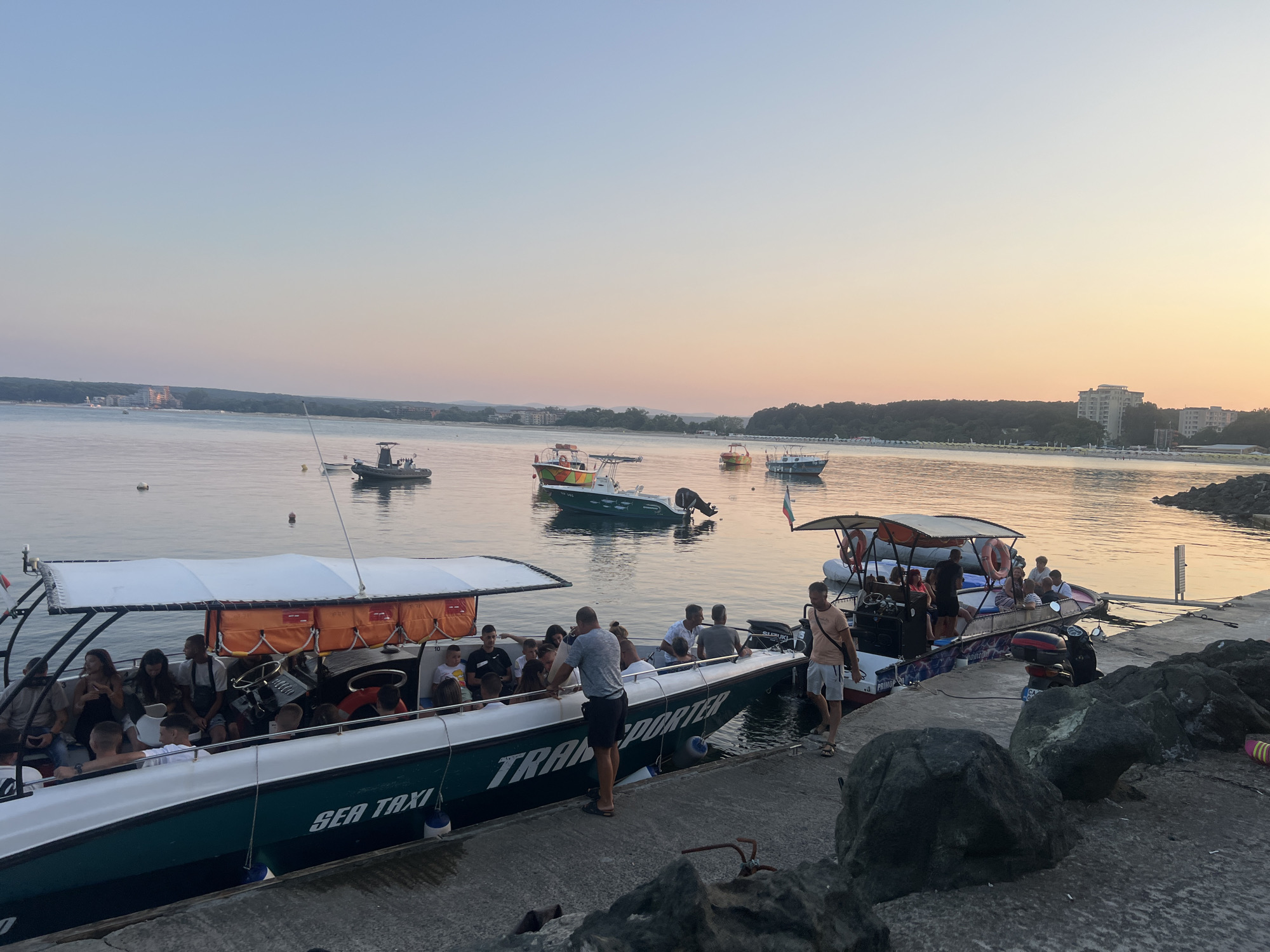 In Primorsko, Bulgaria, at dusk, a group of people are boarding or disembarking from a green and white boat labeled "SEA TAXI TRANSPORTER" docked at a concrete pier. Several other boats are moored in the calm water of a bay. In the background, a tree-lined shore is visible with buildings in the distance. The sky displays a gradient of soft blues and oranges, indicative of sunset. The pier is constructed of concrete and is partially covered with large, dark rocks. A scooter is parked on the pier near the boat.