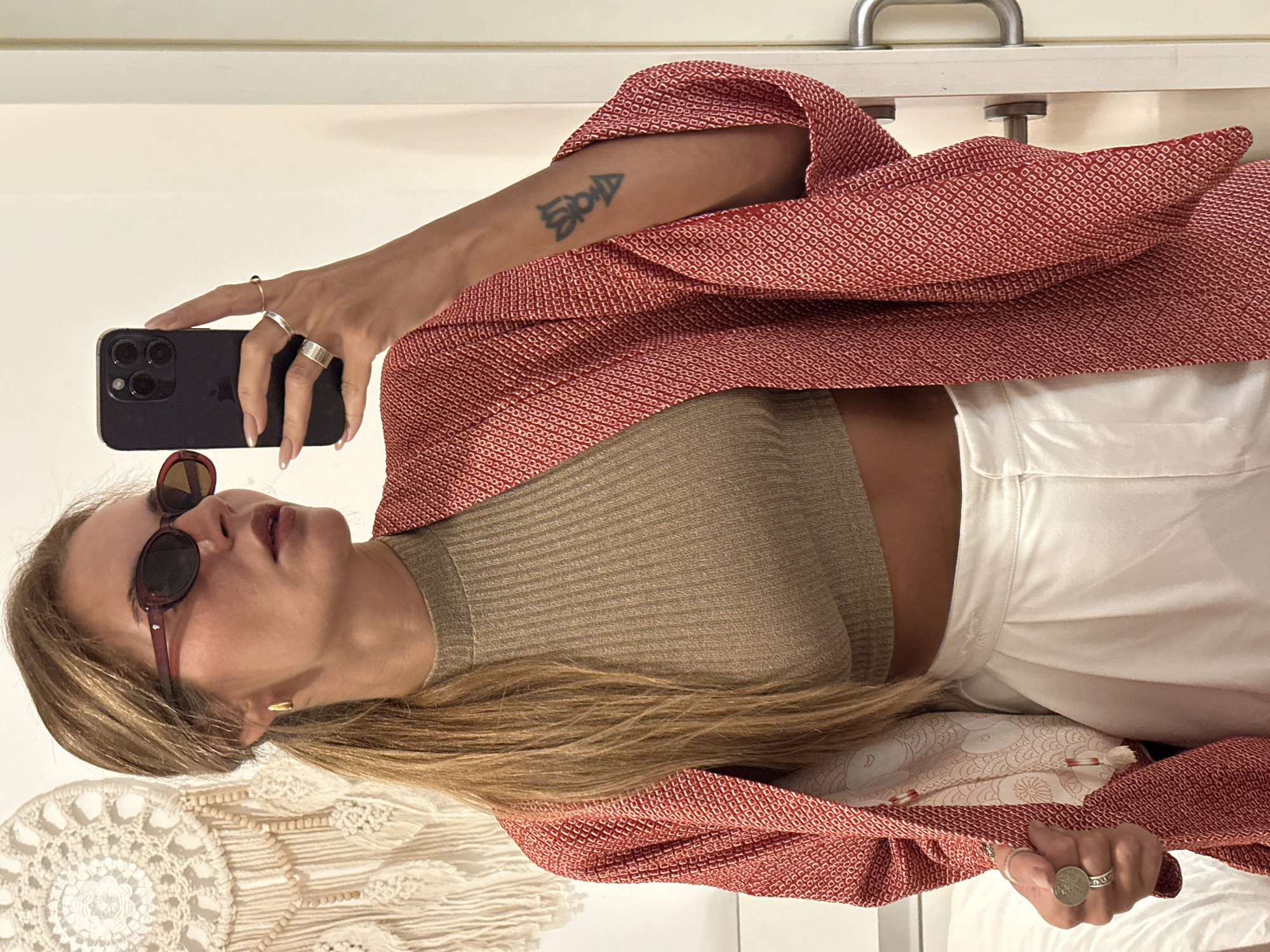 A woman is taking a selfie in what appears to be an indoor setting, possibly a hotel room or apartment. She is wearing sunglasses, a brown knit top, white pants, and a patterned red jacket. She is holding a smartphone in her left hand to take the photo. She has several rings on her fingers. A tattoo is visible on her right arm. The background includes what looks like a headboard, bedding, and a wall. The picture's location is unknown, and there's no explicit indication that this image was taken in Madrid, Spain.