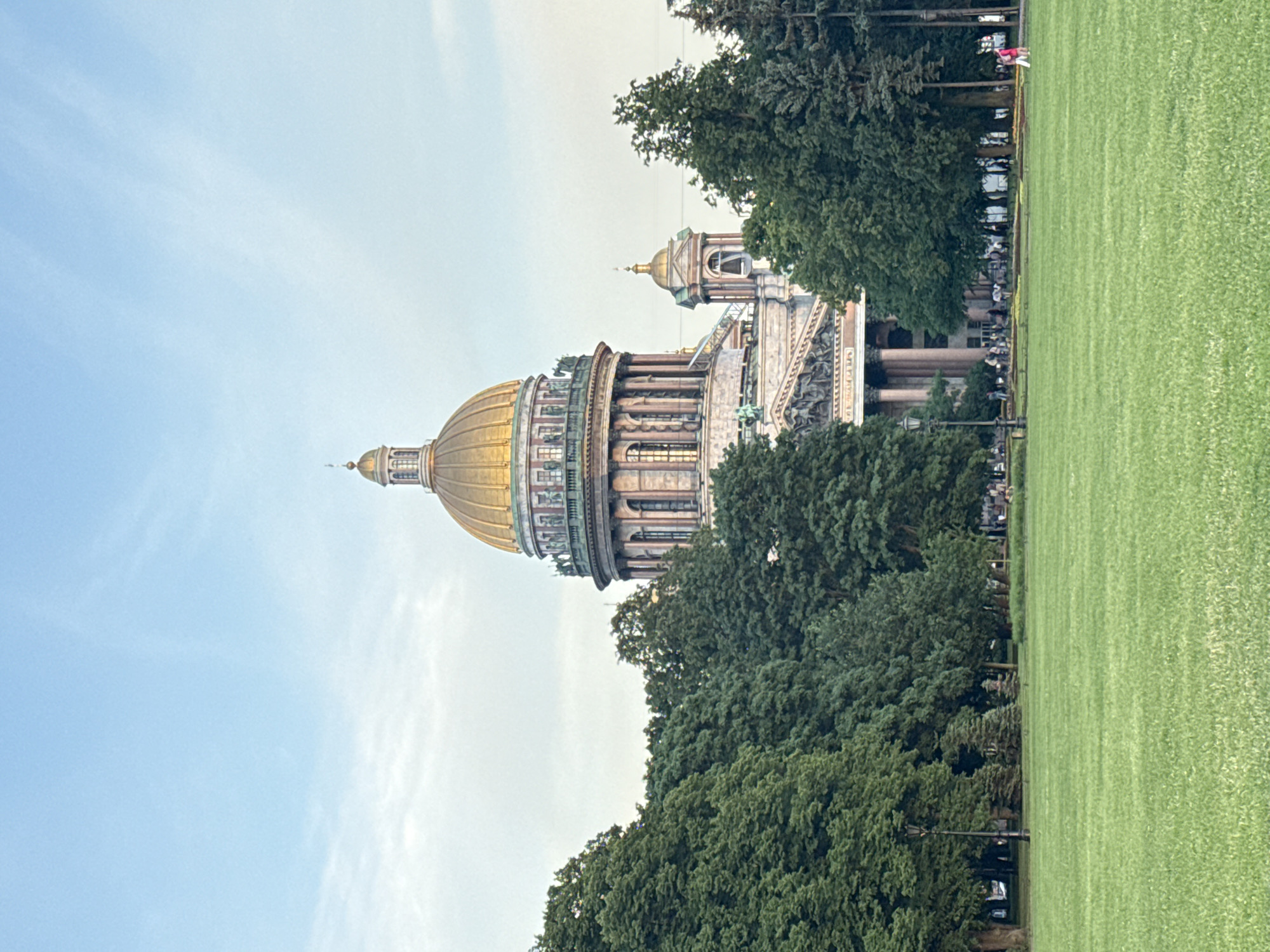 The image shows a view of Saint Isaac's Cathedral in Saint Petersburg, Russia. The cathedral, with its prominent golden dome, is partially visible behind a dense row of lush green trees. In the foreground, a large expanse of well-maintained green grass fills the lower half of the frame. Several people are scattered across the grass and near the cathedral. Some are walking in groups, while others are standing. The sky above is a soft blue with a few wispy clouds. The lighting suggests it is either early morning or late afternoon.
