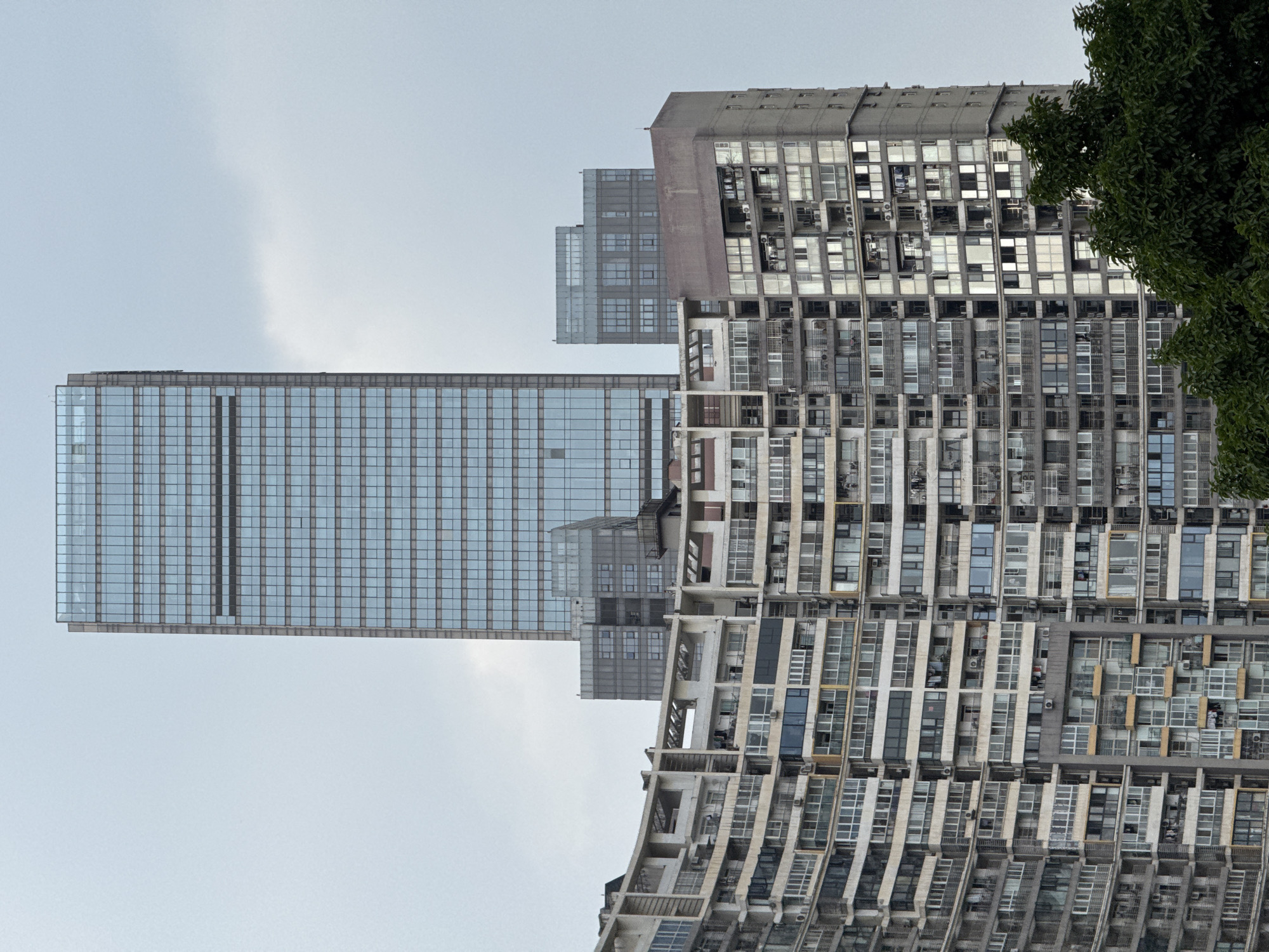 The image presents an urban scene in Wuhou District, China, characterized by a stark contrast between modern and older high-rise architecture under a uniformly overcast sky.
In the background, a very tall, slender, rectangular skyscraper dominates the upper central portion of the frame. Its facade is composed primarily of blue-tinted glass panels arranged in a tight grid pattern, reflecting the grey sky. This structure exemplifies contemporary high-rise design. Directly below and behind the skyscraper, two smaller, boxier buildings with darker grey, grid-patterned facades are visible, suggesting ancillary structures or adjacent developments.
The majority of the foreground is occupied by a complex of densely packed residential apartment buildings. These structures appear older and more weathered, featuring facades in various shades of grey, beige, and white. The prominent blocks on the left exhibit a distinctive curvilinear architectural design, with numerous balconies and windows curving along their extensive length. The buildings are characterized by a multitude of rectangular windows, many of which are enclosed with glass or translucent panels, and an abundance of white air conditioning units affixed to exterior walls and balconies. Visible items on some balconies, such as hanging laundry, indicate active residential occupancy.
The sky above is a uniform light grey, indicative of overcast conditions, providing a muted backdrop for the urban structures. In the bottom right corner of the frame, the dense, dark green foliage of one or more trees partially obscures the base of the residential buildings. No individuals are visibly present in the scene.