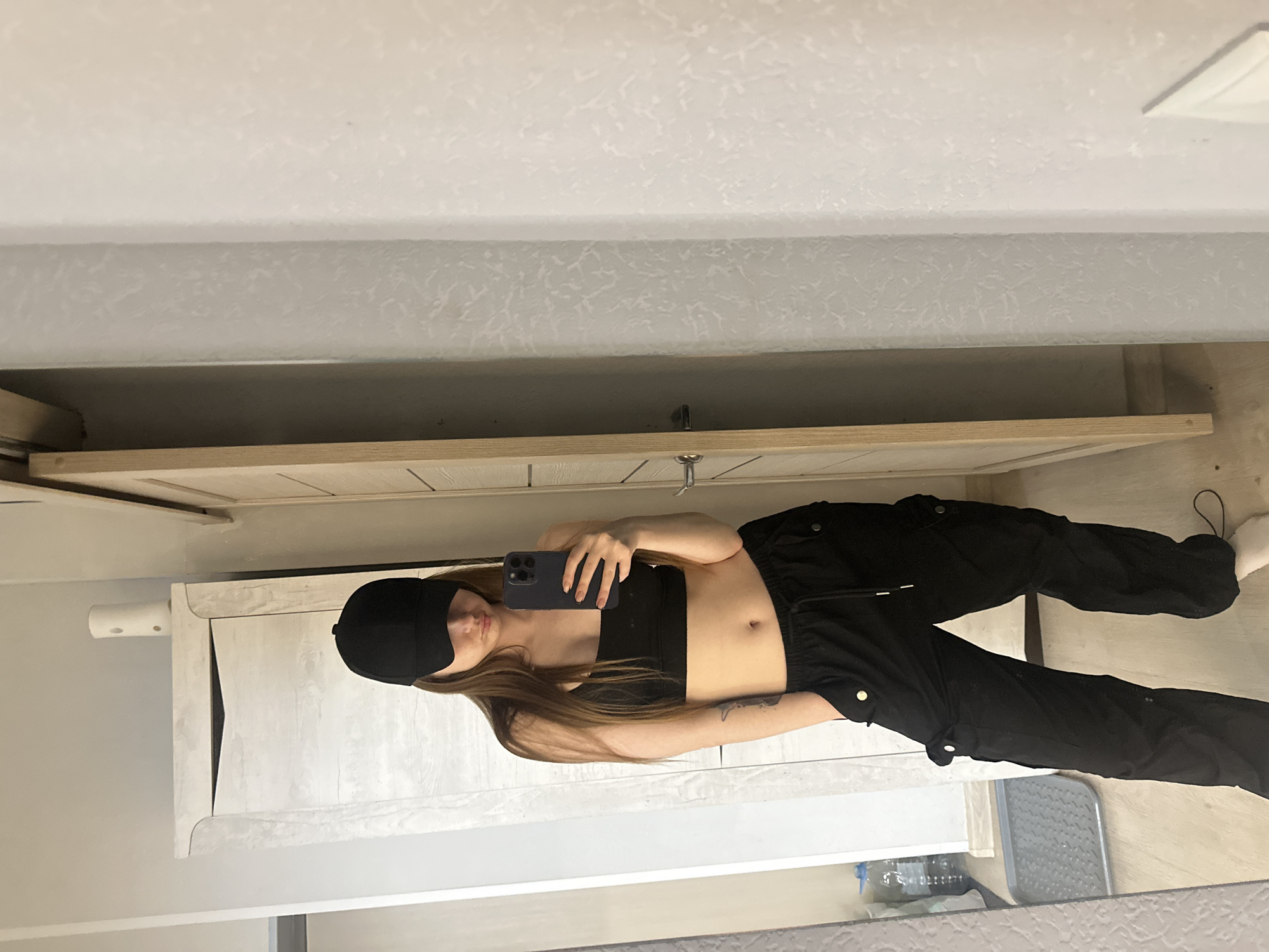 A young woman is standing in an indoor environment. The location appears to be a bathroom, evident by a vanity with drawers and a possible mirror. She is facing the camera, holding a smartphone, apparently taking a selfie. She is wearing a black cropped top, black cargo pants, and a black beanie. Her hair is long and light brown. A tattoo is visible on her arm. The lighting is bright and appears to be natural light from an unknown source. There are several items under the vanity; a water bottle. The city and specific location are unknown.