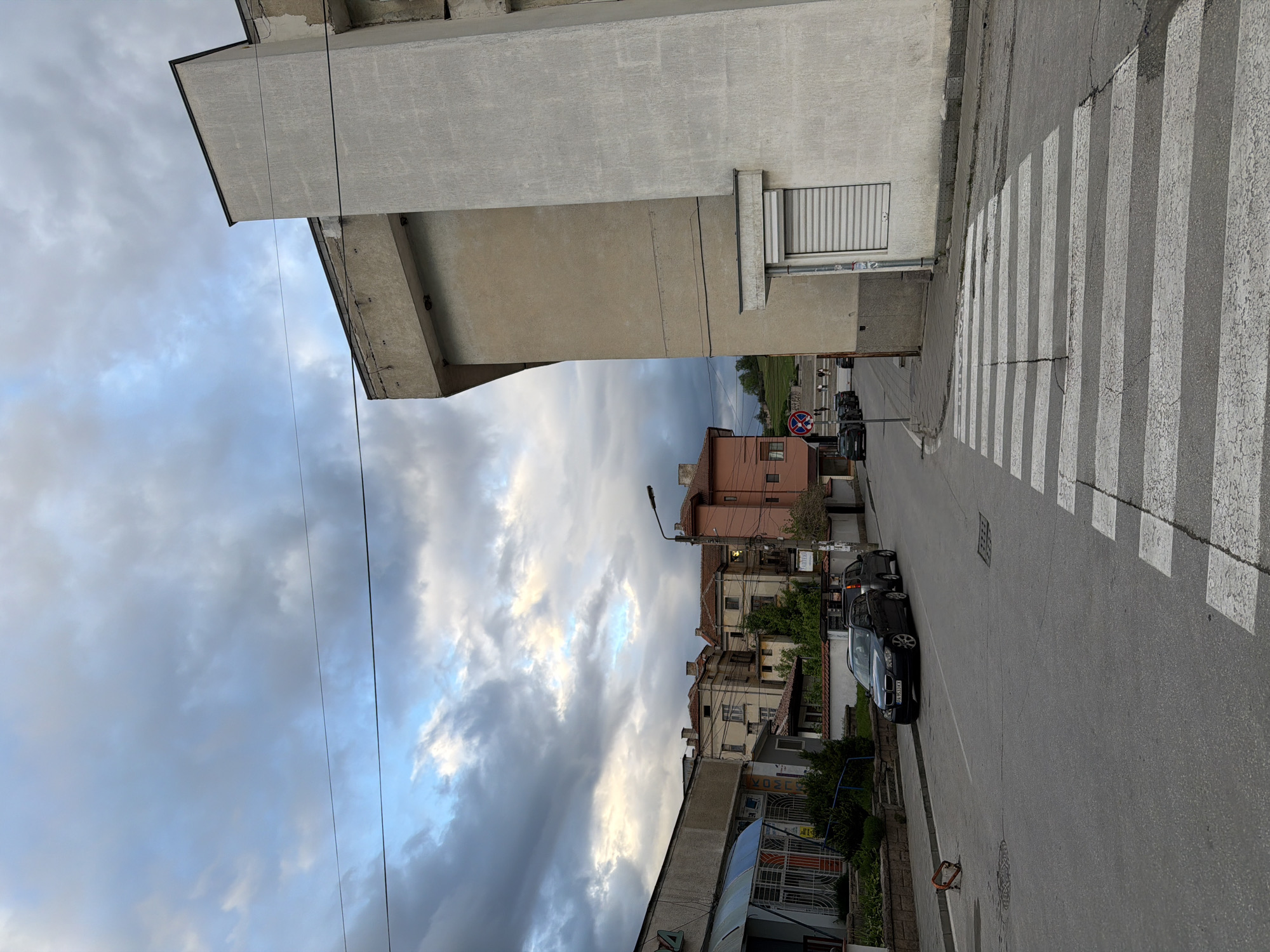 The image shows a street scene in Panagyurishte, Bulgaria. The street is paved with asphalt and features a crosswalk with white stripes. Buildings of varying heights and architectural styles line both sides of the street. A dark-colored car is parked on the right side of the street, and additional vehicles are visible further down the road. A street lamp and several utility poles are present. The sky is filled with a mix of light and dark clouds, suggesting either an approaching storm or clearing weather. A no parking sign is visible. The buildings appear to be a mix of residential and commercial properties. The image is slightly rotated counter-clockwise.