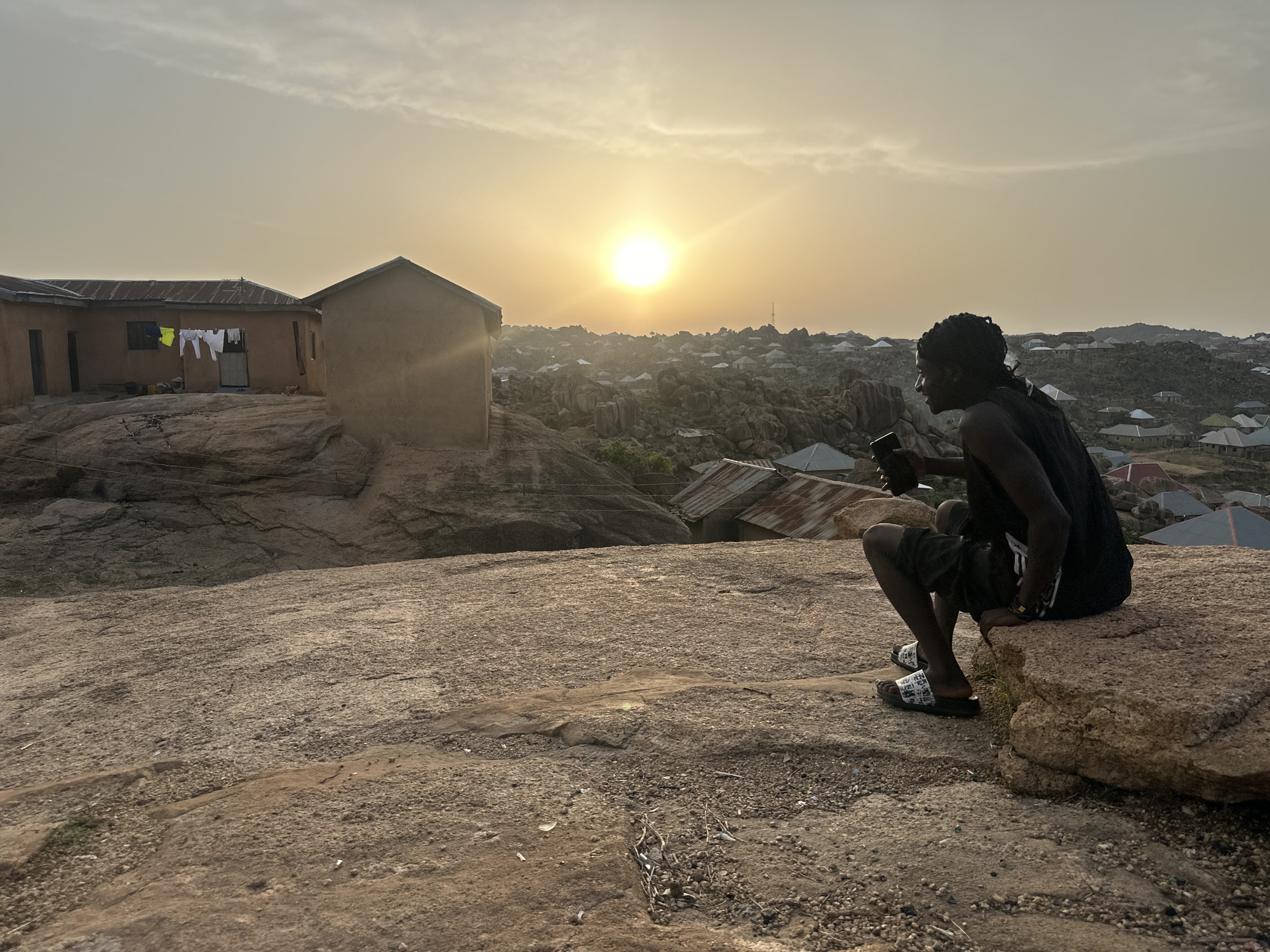 A person with dark skin and braided hair sits on a rock overlooking a settlement in Jos, Nigeria. The individual is wearing a black tank top, dark shorts, and patterned sandals. They are holding a smartphone or similar device and looking towards the setting sun. In the background, there are houses constructed on and around rocky outcrops, with laundry hanging from one of the buildings. The landscape consists of bare rock formations and scattered dwellings, under a sky with the sun low on the horizon and light cloud cover.